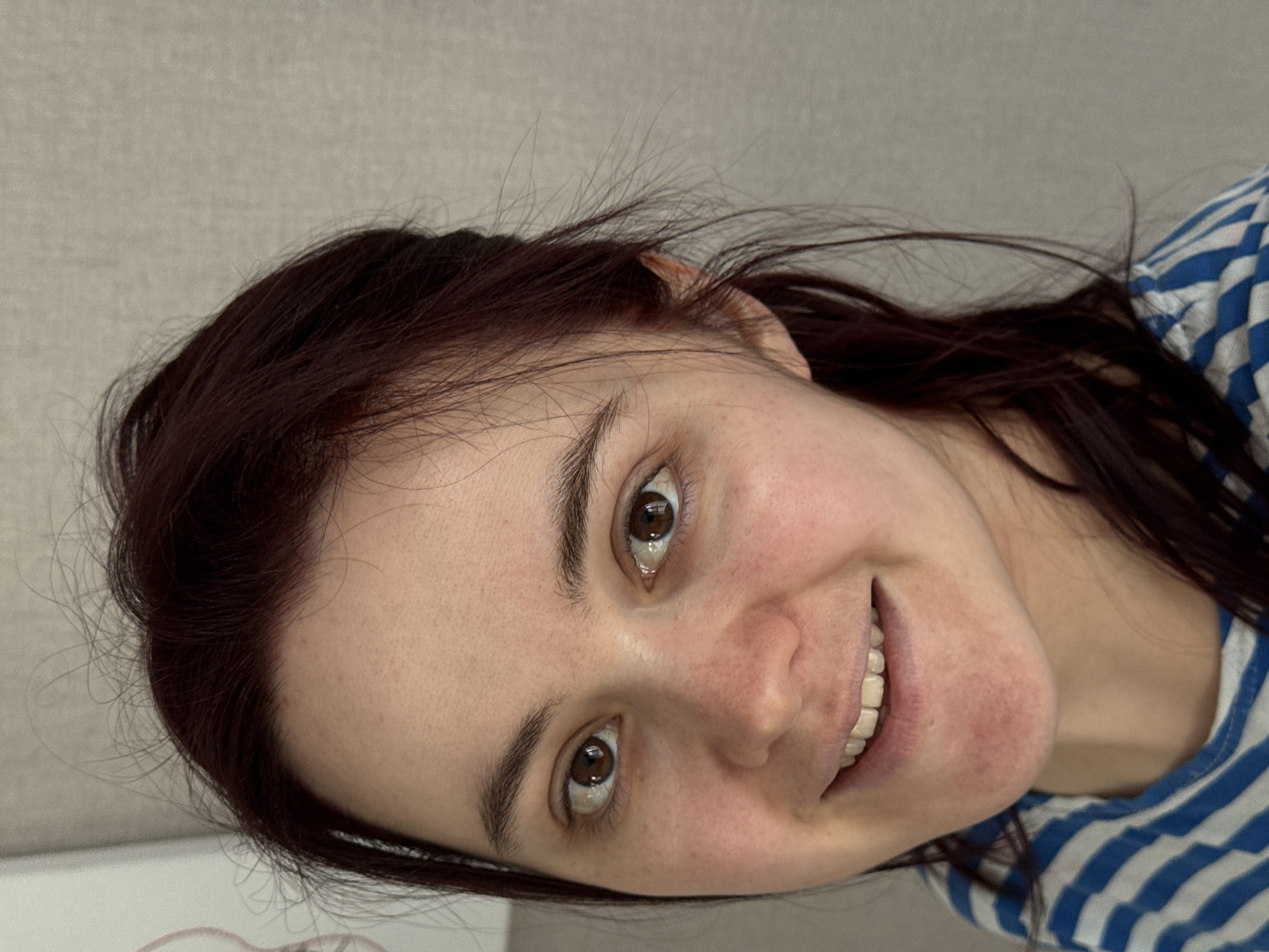 A woman with dark red hair and brown eyes is smiling at the camera. She has fair skin with freckles. She is wearing a blue and white striped shirt. The background is a plain, light-colored wall. The location is not identifiable as Izhevsk, Russia.