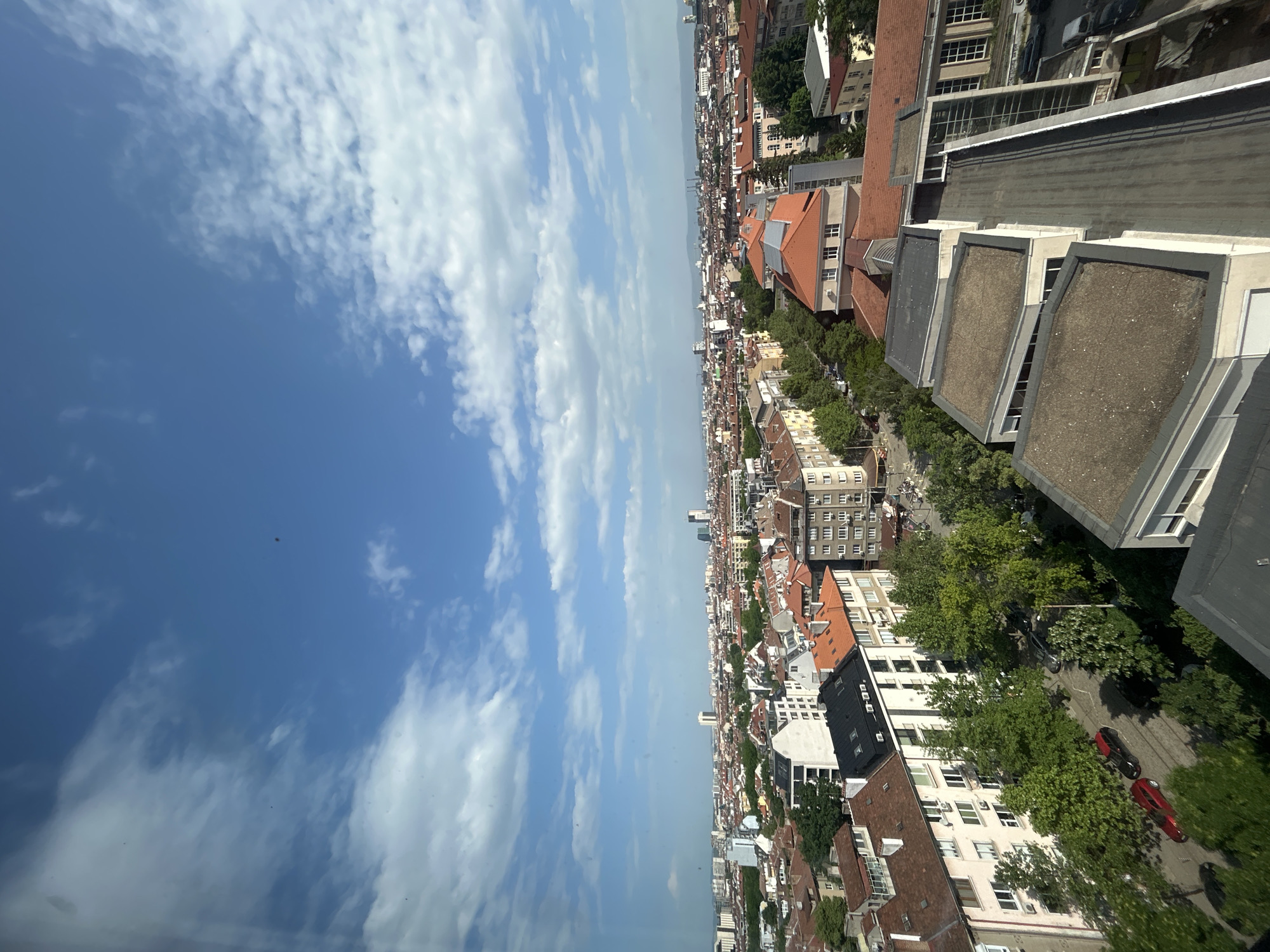 Here's a detailed description of the image:
The photograph presents an aerial view of Sofia, Bulgaria, under a partially cloudy sky. The focus is on the urban landscape, predominantly featuring buildings with a mix of red-tiled and flat, concrete roofs. The building styles vary, indicating different periods of construction within the city.
Greenery is visible in the form of trees lining the streets and interspersed between buildings, suggesting planned urban forestry. Several cars are parked or moving along streets that weave through the buildings.
The sky occupies approximately half of the image, exhibiting a bright blue color punctuated by wispy, elongated clouds.
No people are clearly visible in the image, as the perspective is too high to distinguish individual figures. The city appears active due to the presence of cars and general urban planning, but lacks clear indicators of significant activity in any particular area.
The photograph seems to have been taken from a high vantage point, possibly from a skyscraper or a tall communication tower. The quality of the image is clear, allowing for the identification of the distinct architectural styles and features of Sofia's urban environment.