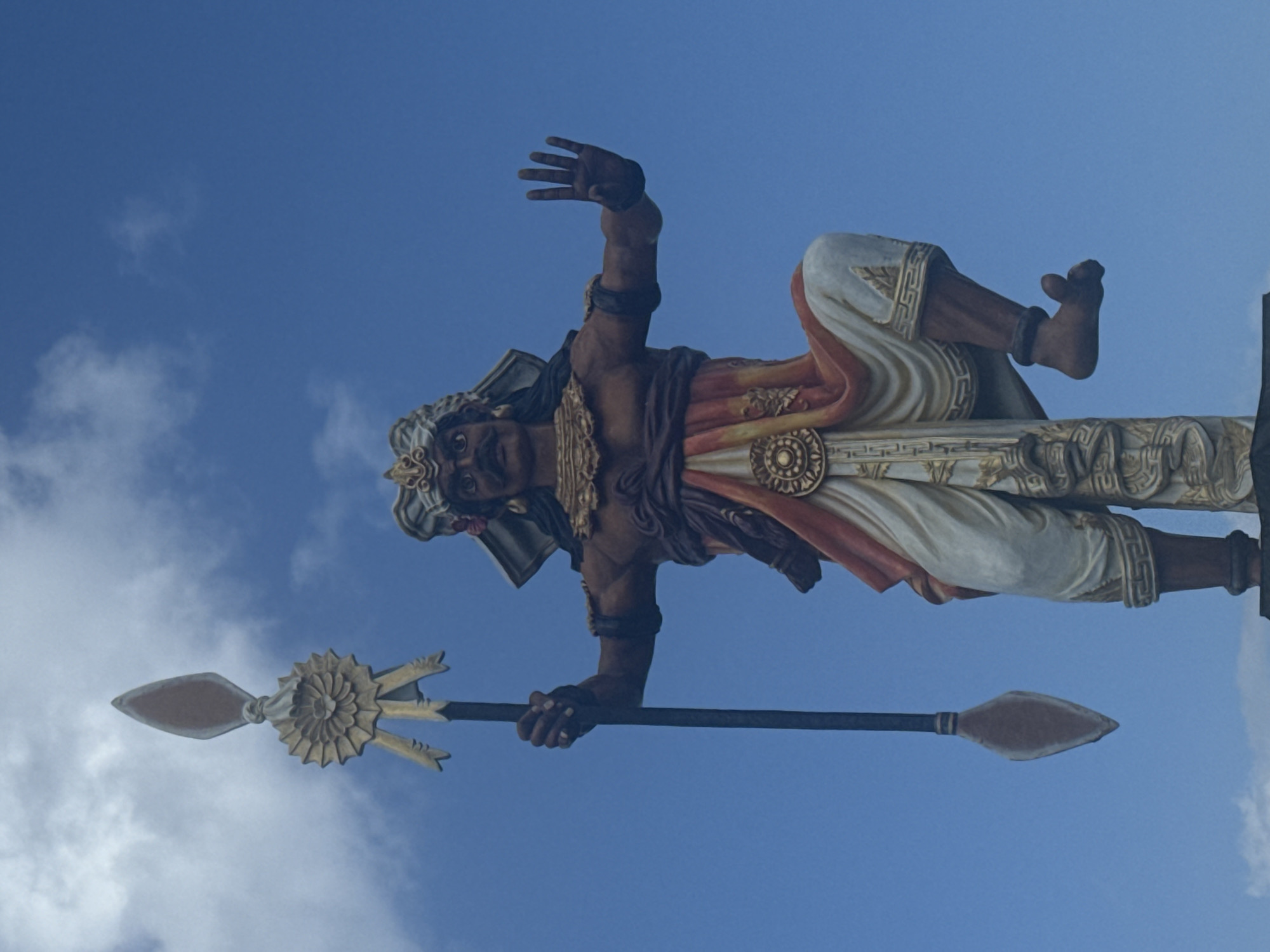 A full-body, low-angle shot shows a colorful, large statue of a warrior or deity against a clear blue sky with scattered white clouds. The statue is depicted in a dynamic pose, kneeling on its left leg with its right leg extended forward. It holds a tall spear vertically in its left hand, with the spear's decorative top featuring a sunburst design and ribbons. The statue's attire includes a white and orange draped garment around its waist and legs, a patterned band around its calves, and dark armbands. It wears an ornate headdress and has a mustache. Its right hand is raised forward with fingers spread, as if warding off something or making a gesture. The statue appears to be made of painted stone or fiberglass. The background is an expansive, cloud-filled sky, suggesting an outdoor location. The statue is the sole subject in the frame.