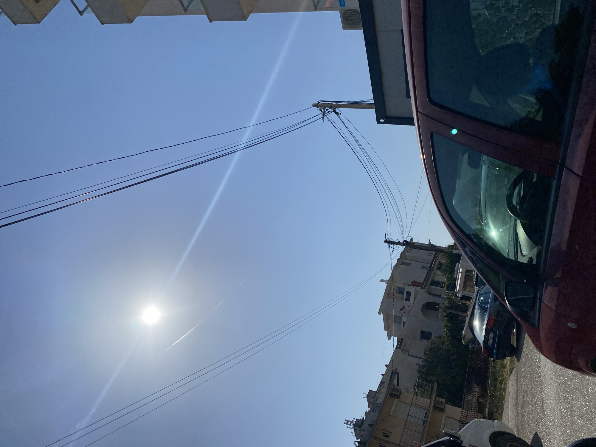 This image captures a sunlit street scene in Sutomore, Montenegro, with a clear blue sky dominating the upper portion. The sun is bright and high, creating lens flare and casting light across the scene. Overhead, a series of dark power lines crisscross the sky, supported by wooden utility poles.
In the foreground, the burgundy roof and partial side view of a car are visible, with its windows reflecting the surroundings. To the left, a white van is partially in frame. Behind the vehicles, a row of buildings lines the street. These buildings appear to be residential, with multiple stories, balconies, and visible air conditioning units. Some of the buildings have a light-colored facade, while others are darker. Vegetation, including trees, is interspersed between the buildings.
Further down the street, several other cars are parked or driving. One dark sedan with a license plate visible (NJ V333 LR) is positioned in the middle ground, its surface reflecting the sunlight. The road surface appears to be asphalt. The overall impression is that of a typical urban or semi-urban environment on a clear, sunny day.