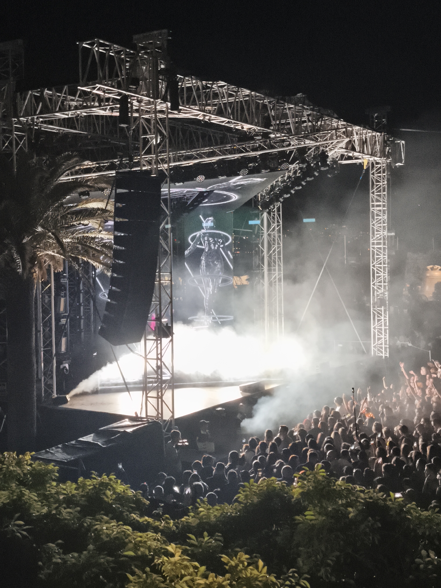 The image shows a concert at night, likely outdoors. A crowd of people is visible in the foreground, many with their arms raised, suggesting they are enjoying the performance. A stage is set up in the background, with metal scaffolding supporting lights and speakers. A holographic figure is displayed on the stage. Fog or smoke is present, partially obscuring the view. Some foliage, possibly trees or bushes, is visible in the lower foreground. The location is identified as Unknown, Unknown.