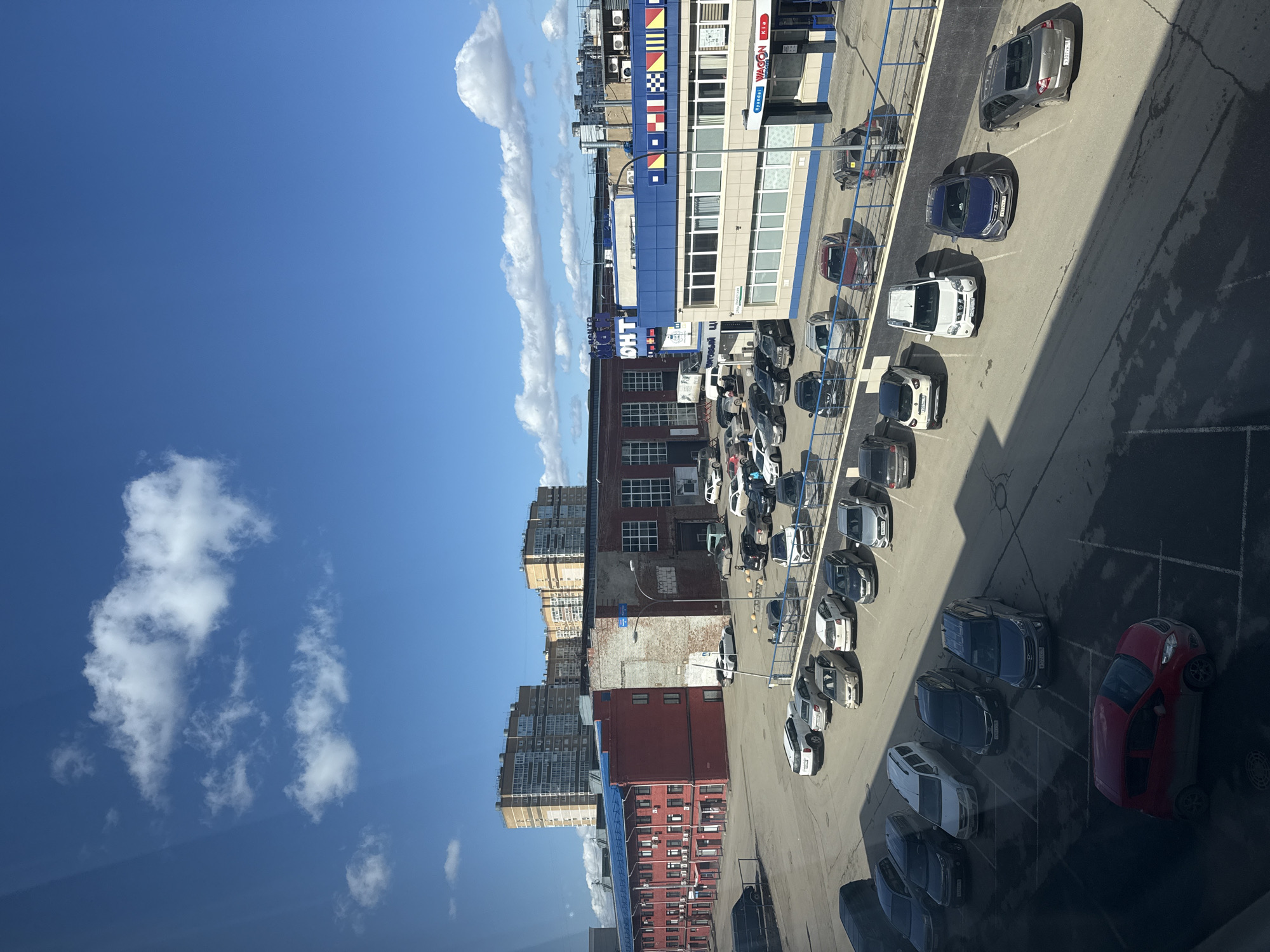 The image shows an aerial view of a parking lot adjacent to buildings in what appears to be Izhevsk, Russia. Clear blue sky with scattered clouds is visible. A multistory building with blue and white facade, displaying nautical flag designs, can be seen. "Wagon Kia Hyundai" is on the building, written in English. Adjacent to the building are older brick structures of varying heights, typical of urban environments. The parking lot contains numerous parked cars, most of which are sedans, in shades of white, gray, blue, and red. The parking lot pavement shows signs of wear and has cracked pavement. A tall residential building is in the background. Shadows cast by the surrounding buildings are visible on the right side of the parking lot.