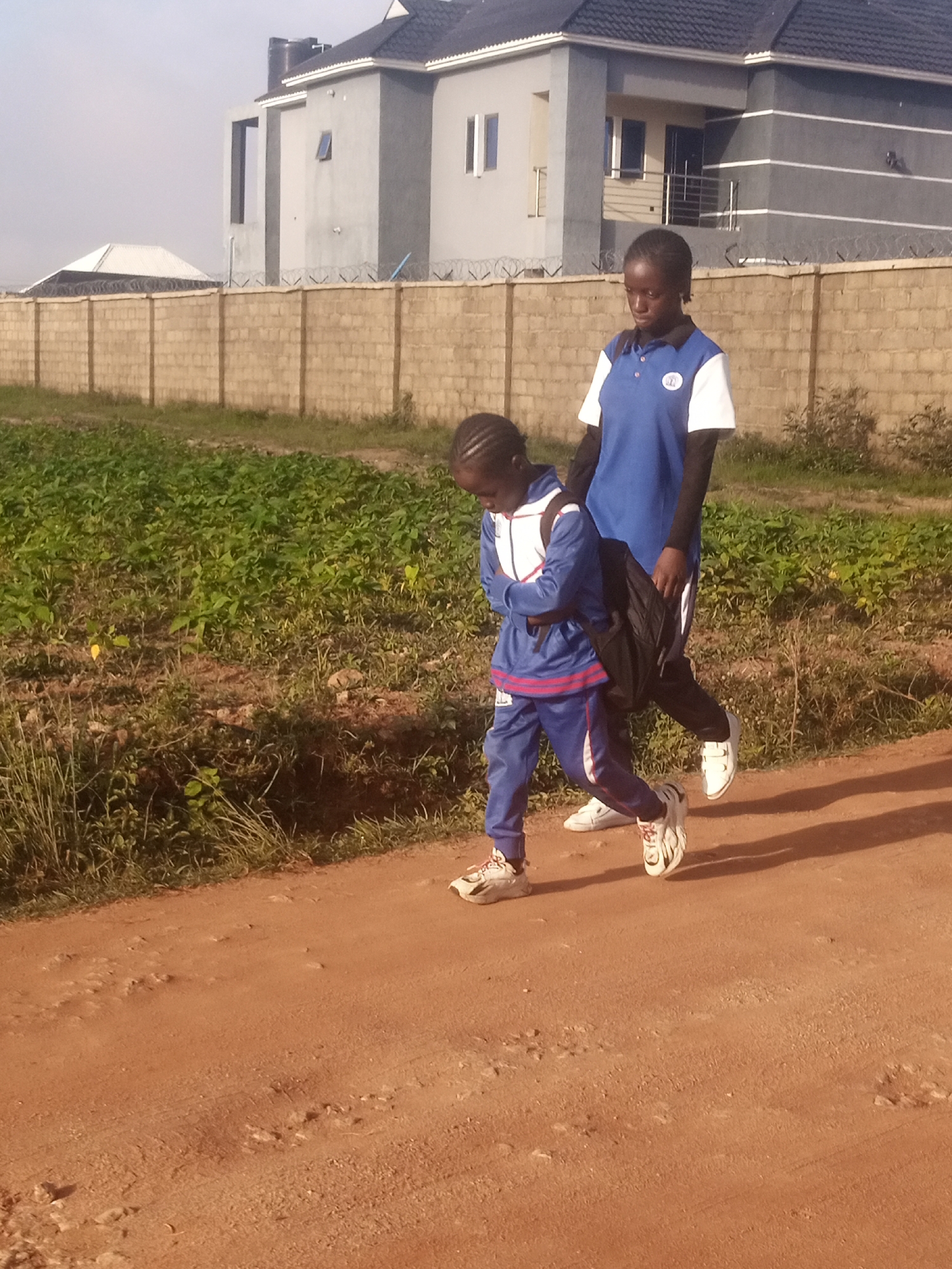 Two young females are walking on a dirt road. The female in the foreground is wearing a blue and red tracksuit and carrying a black backpack. She is looking down and appears to be walking. The female behind her is wearing a blue and white polo shirt and black pants. She is looking forward and appears to be walking as well. They are walking past a field of green plants and a brick wall. In the background, there is a large, modern-looking building with a grey exterior and a dark roof. The sky is clear and bright.