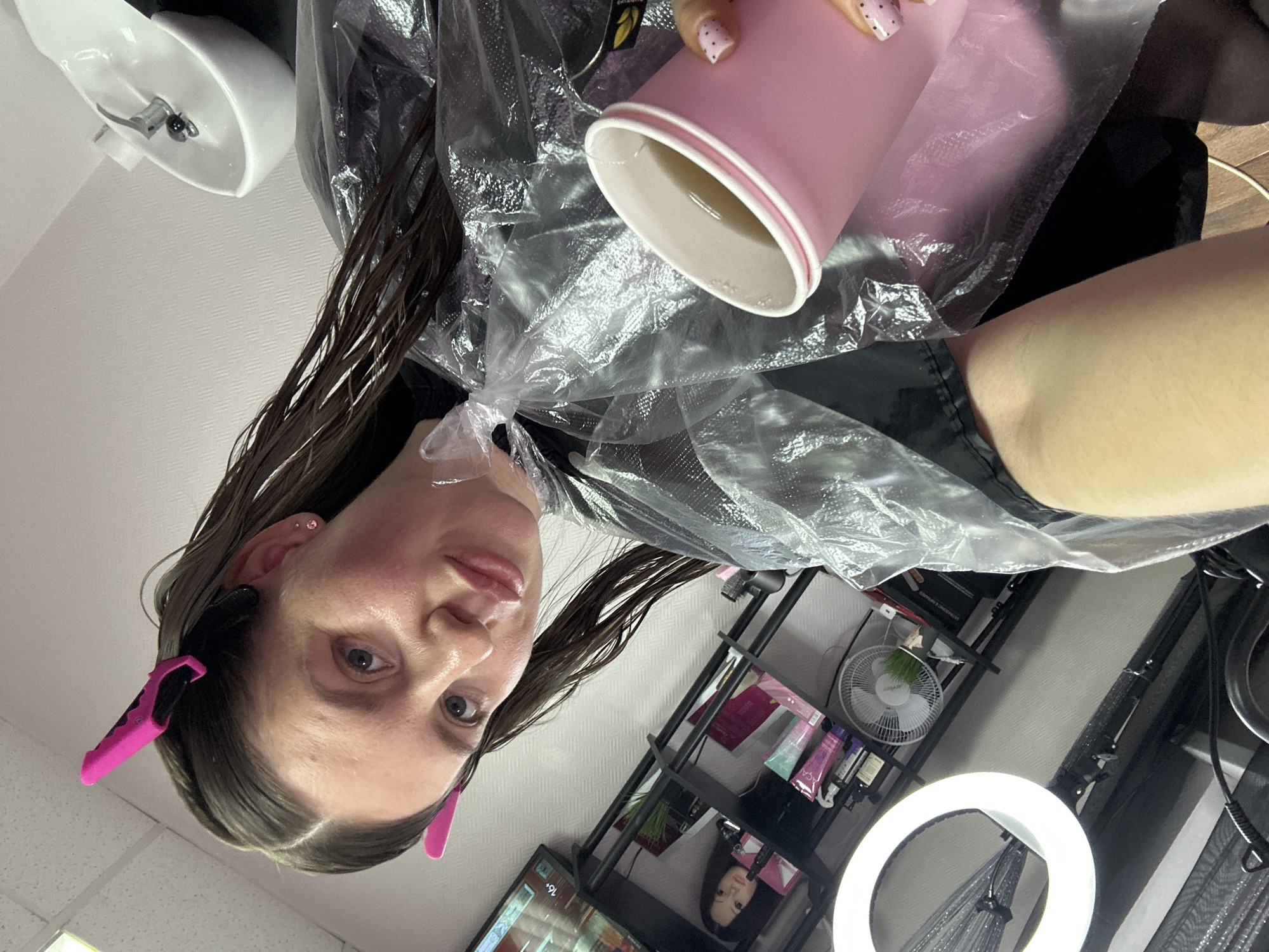 A woman is pictured indoors, presumably in Klin, Russia, based on visual indicators like the Cyrillic script on packaging. She is wearing a plastic hair-dyeing cape and pink hair clips. She is holding a pink and white cup containing a light-colored liquid. In the background, there's a shelf unit containing various beauty products, a small fan, and a ring light. Additionally, a hair salon sink is visible in the top left corner of the image.