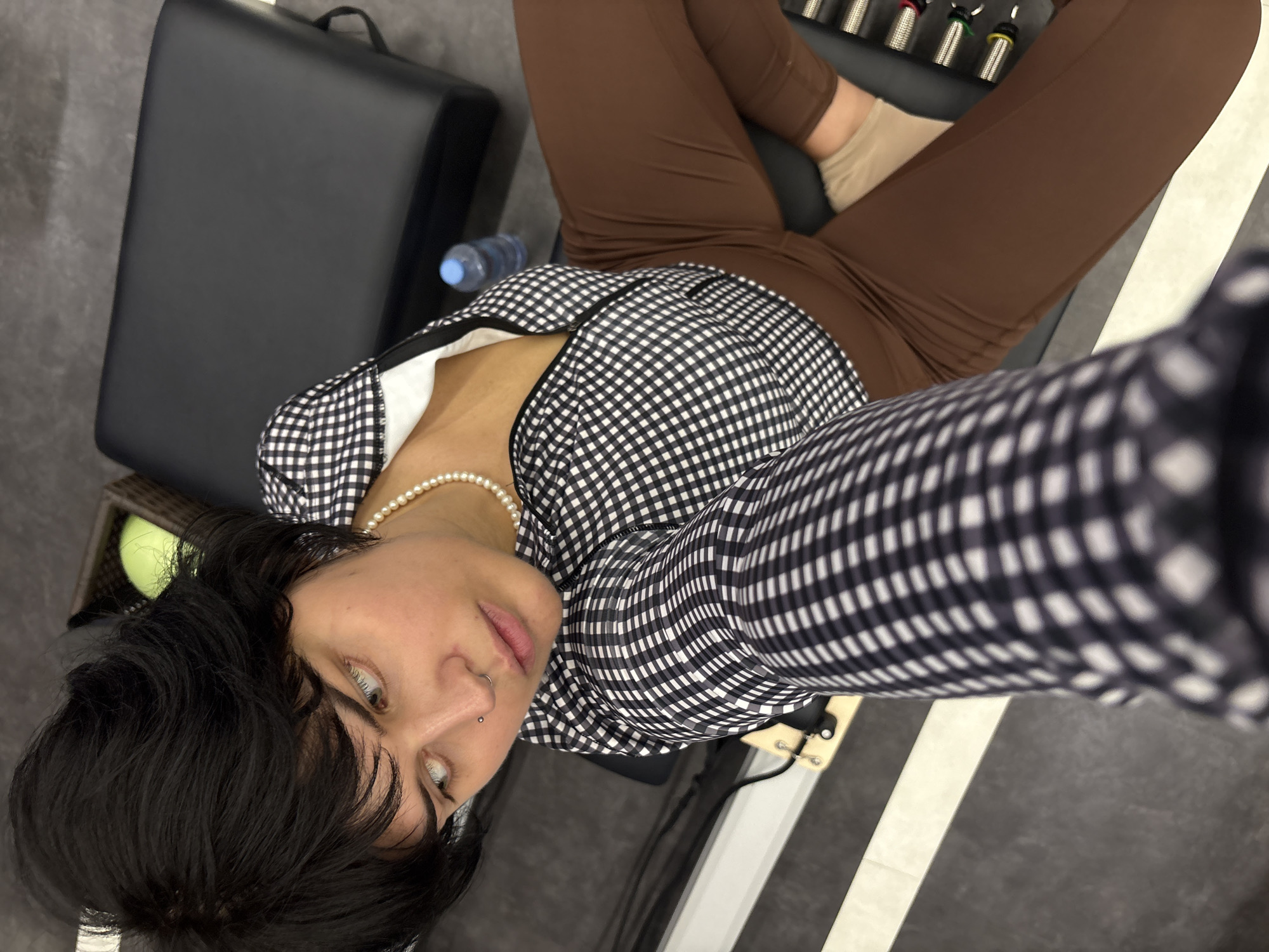 The image shows a person sitting on a piece of exercise equipment. The person has short, dark hair, light-colored eyes, and a nose ring. They are wearing a black and white checkered zip-up jacket over a white top, brown leggings, and light-colored socks. A pearl necklace is visible around their neck. Behind the person, a black exercise machine with springs is visible. To the left of the machine, a basket containing a green object is present. A water bottle is also visible on the machine. The floor appears to be dark gray with a lighter colored stripe. The scene is indoors and appears to be a fitness or exercise studio. There is no indication of the location being in Nong Pa Khrang, Thailand.