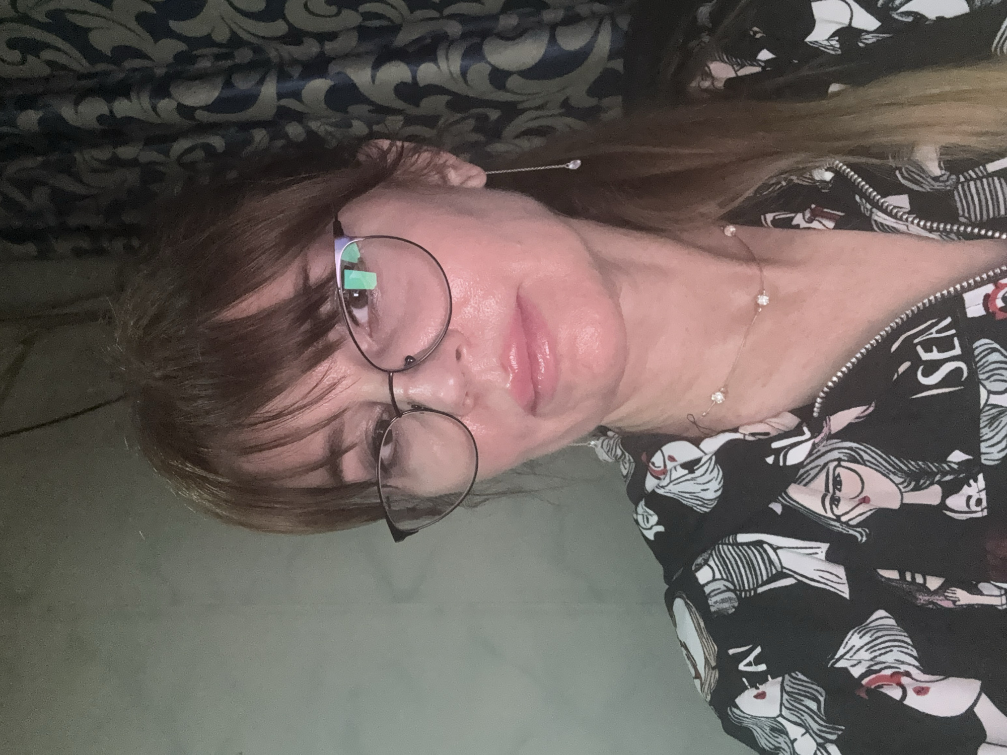 A young woman with fair skin, wearing round glasses and a black and white patterned zip-up jacket, is lying on a light-colored surface. She has brown bangs and longer blonde hair. She is wearing a delicate necklace and an earring with a fine chain. The background includes a dark blue and silver patterned fabric. The location is not discernible.