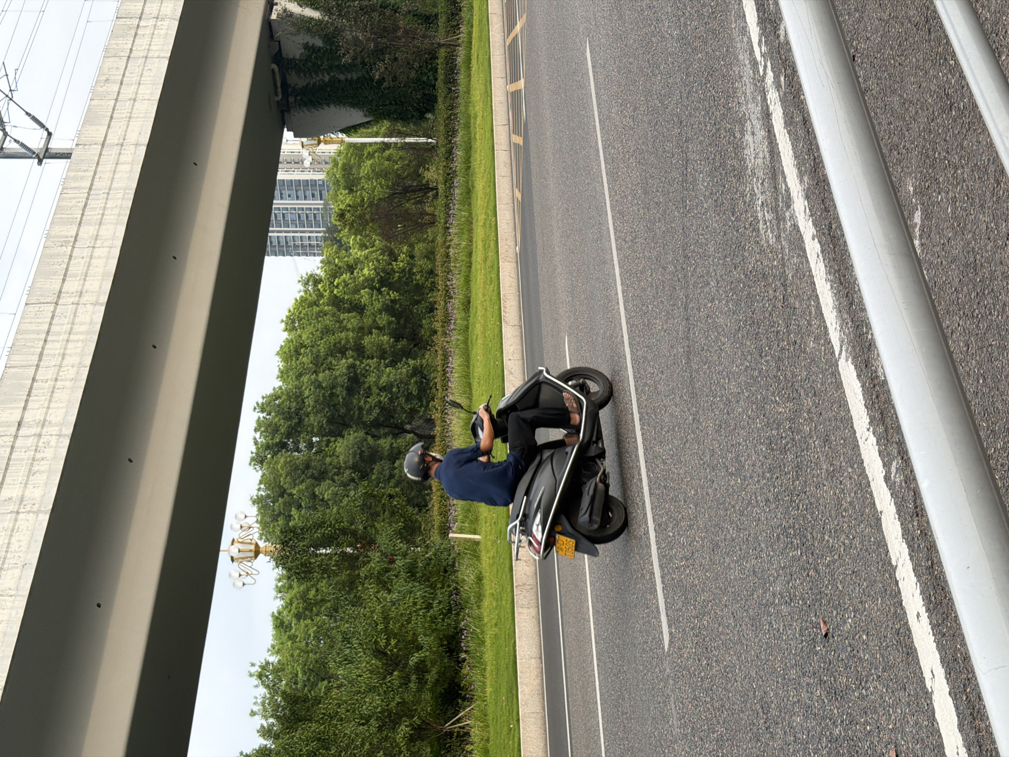 In Jiangjin, China, a person wearing a dark blue shirt, dark pants, and a helmet rides a dark-colored scooter on an asphalt road. The scooter has a yellow license plate. The road has white lane markings. In the background, there are trees, a pedestrian walkway, and a tall concrete overpass with overhead electrical wires for a train. Buildings are visible beyond the trees. The sky is overcast.