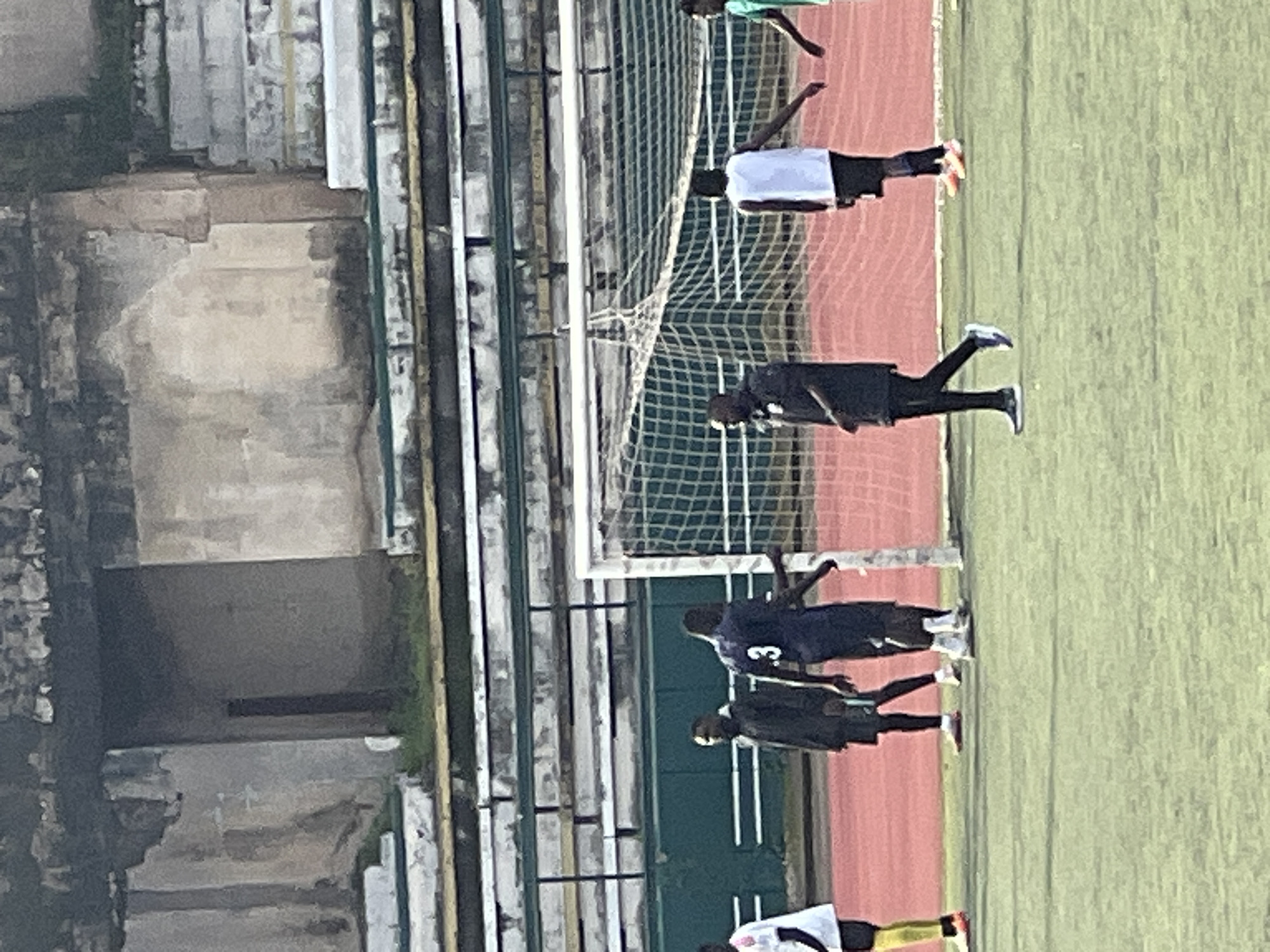 The image depicts a football field at the New Stadium in Nigeria. Several individuals, presumably players, are visible on the field, some near a soccer goal. The stadium appears to be in a state of disrepair, with crumbling concrete structures and weathered seating areas. The overall atmosphere suggests a training session or a match taking place in a somewhat neglected sports facility.
