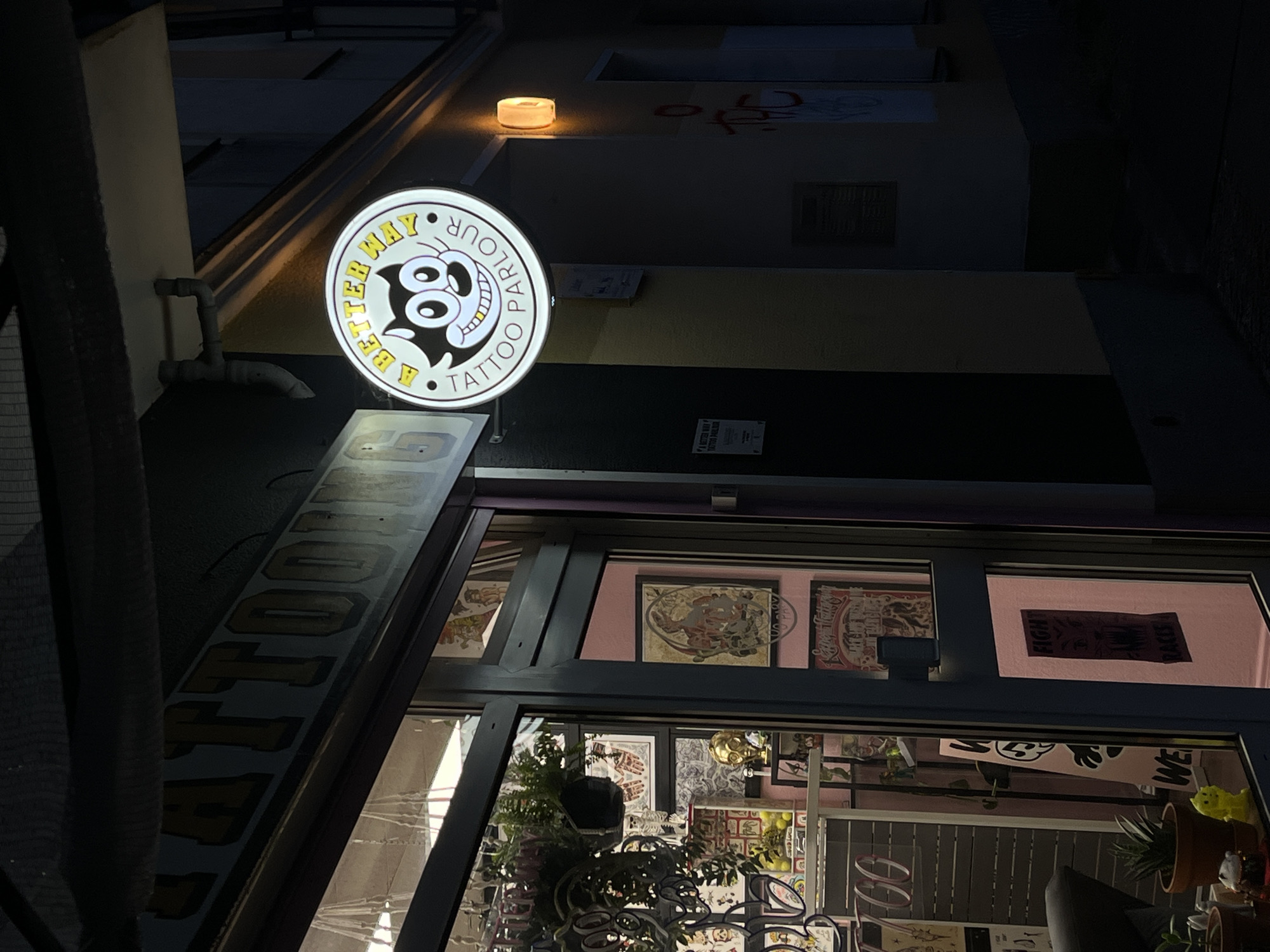 This is a nighttime, medium-shot, eye-level photograph taken outside a tattoo parlor in Berlin, Germany. The establishment's name, "A Better Way Tattoo Parlour," is displayed on a circular, illuminated sign featuring a cartoon cat's face. Above the entrance, a longer sign with yellow, block lettering reads "TATTOOING." The facade of the building is dark, with the interior of the shop illuminated and visible through the large glass windows. The interior displays various tattoo art on the walls and shelves with decorative items, including plants and figurines. A dark-colored door is visible on the right side of the storefront. The street in front of the parlor is dimly lit, with graffiti visible on the adjacent wall.