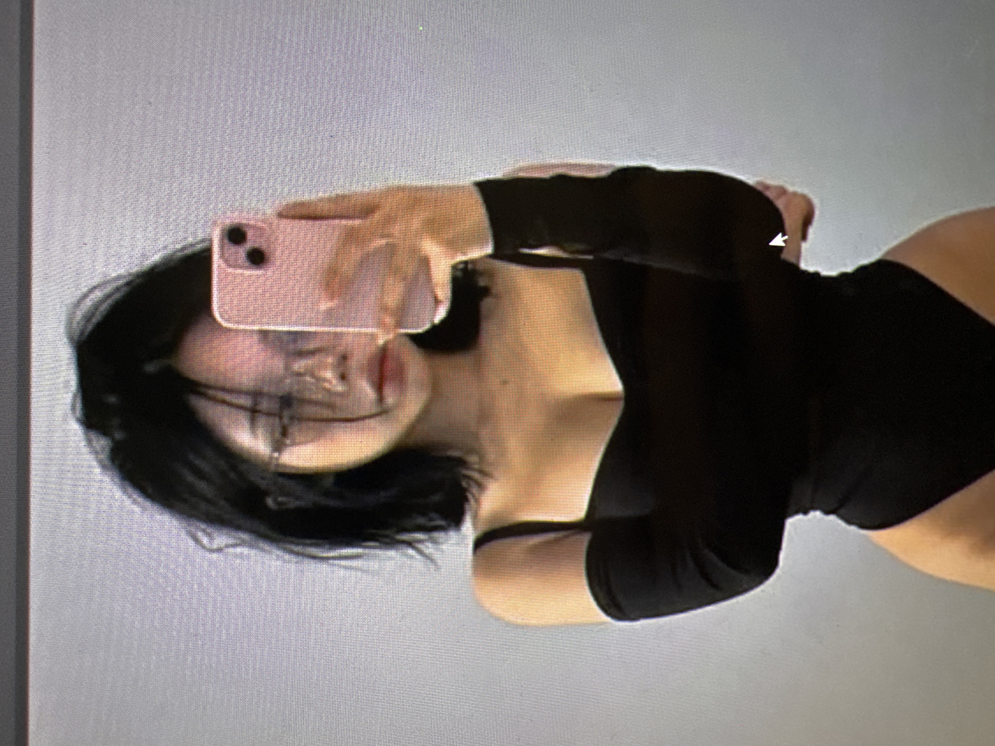 The image is a medium shot of a young woman with dark hair and glasses taking a selfie with a pink smartphone. She is wearing a black off-the-shoulder top with long sleeves. The background is a plain, light-colored wall. The image quality is low, with visible pixels. There is no information available about the location being Castelo Branco, Portugal.