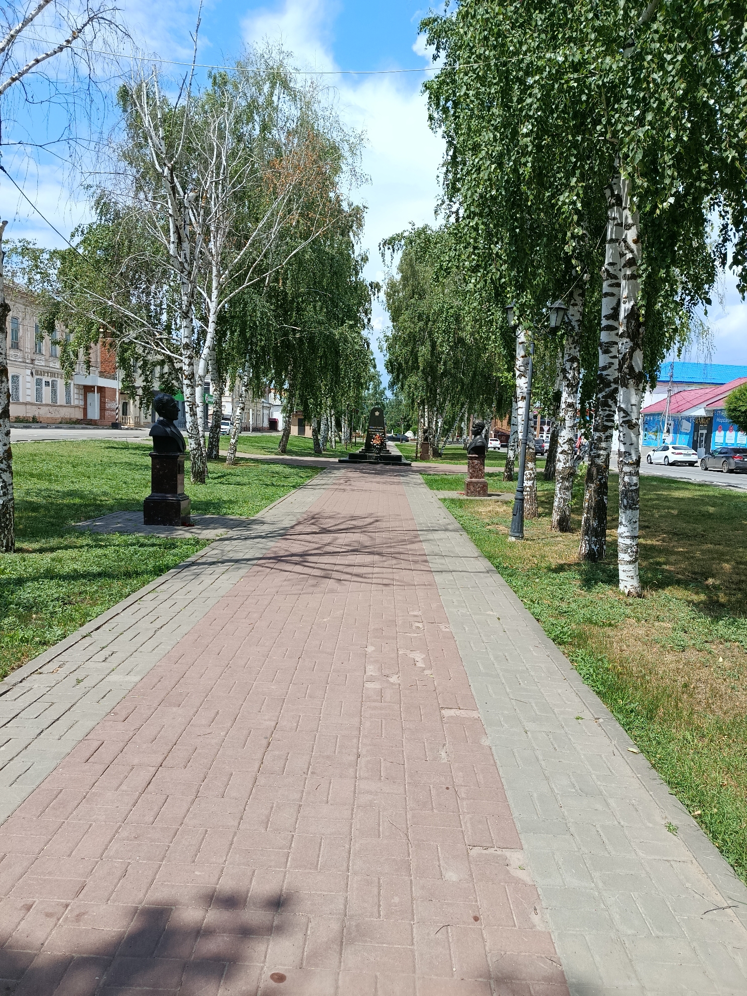 The image depicts a tree-lined pedestrian walkway in the city of Marx, Russia. The walkway is paved with reddish-brown and grey interlocking bricks, leading towards a monument at the end of the path. On either side of the walkway are grassy areas with scattered trees, predominantly birch trees with their distinctive white bark.
To the left of the walkway, a building with multiple windows and architectural detailing is visible behind the trees. Further down the path on the left, a bust is mounted on a dark pedestal. On the right side of the walkway, more birch trees are present, with a street visible beyond them. Several cars are parked along the street, and a building with a blue roof and signs is also visible.
In the distance, at the end of the walkway, a memorial monument with what appears to be a red flame or light can be seen. The sky is blue with scattered white clouds, suggesting fair weather. There are no people clearly visible in the immediate foreground, though shadows on the walkway indicate potential presence or past activity. The scene conveys a sense of public space, likely a park or a memorial avenue within the city.