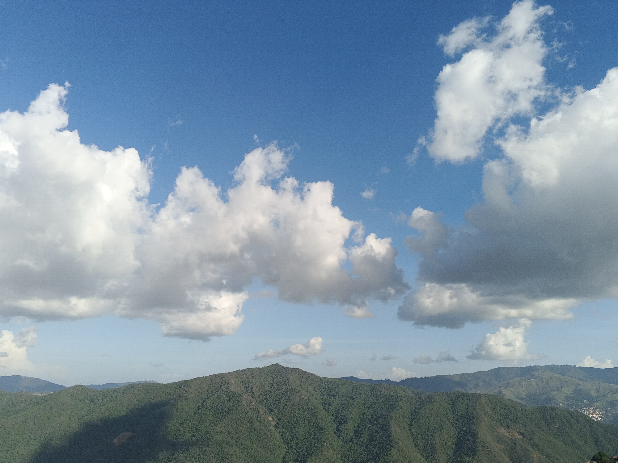 The image shows a landscape featuring green, forested mountains under a bright blue sky dotted with white cumulus clouds. The mountains stretch across the horizon, with varying heights and densities of vegetation. The sky occupies the majority of the frame, with the clouds scattered across its upper portion. The scene depicts a natural environment. This location is most likely to be from Caracas, Venezuela.