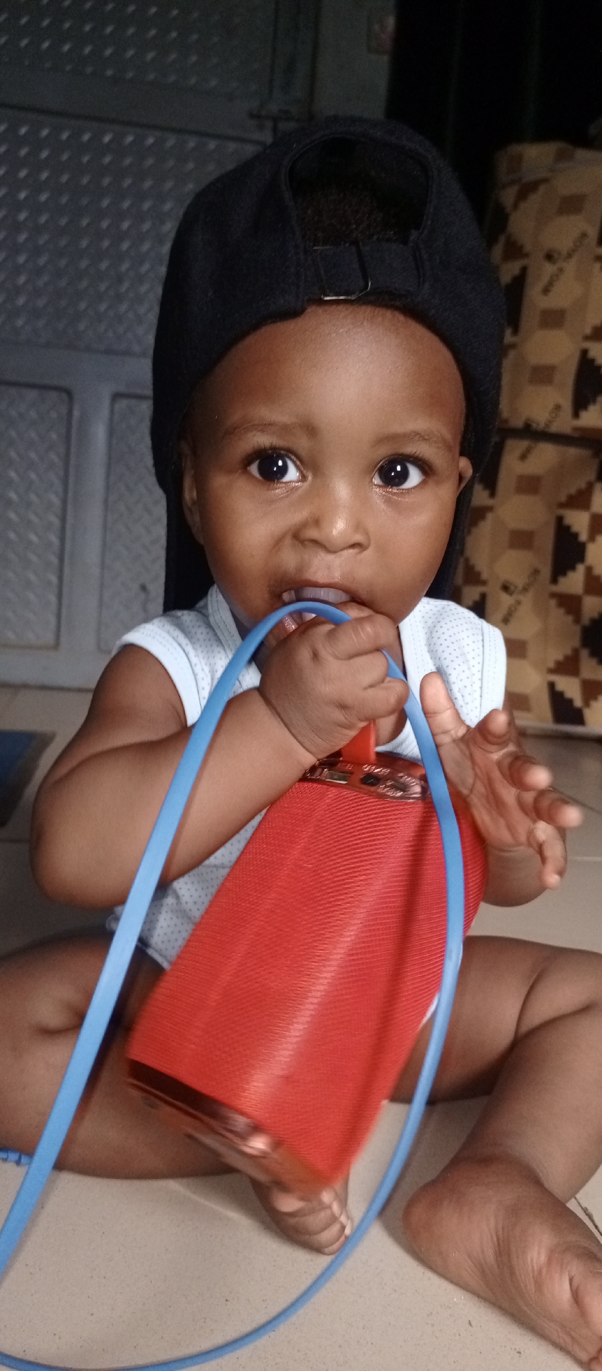 A baby wearing a black cap sits on the floor. The child is holding a red speaker with a blue strap and has the strap in their mouth. The location is Unknown, Unknown. The background includes a textured metal panel and a surface with a geometric pattern.
