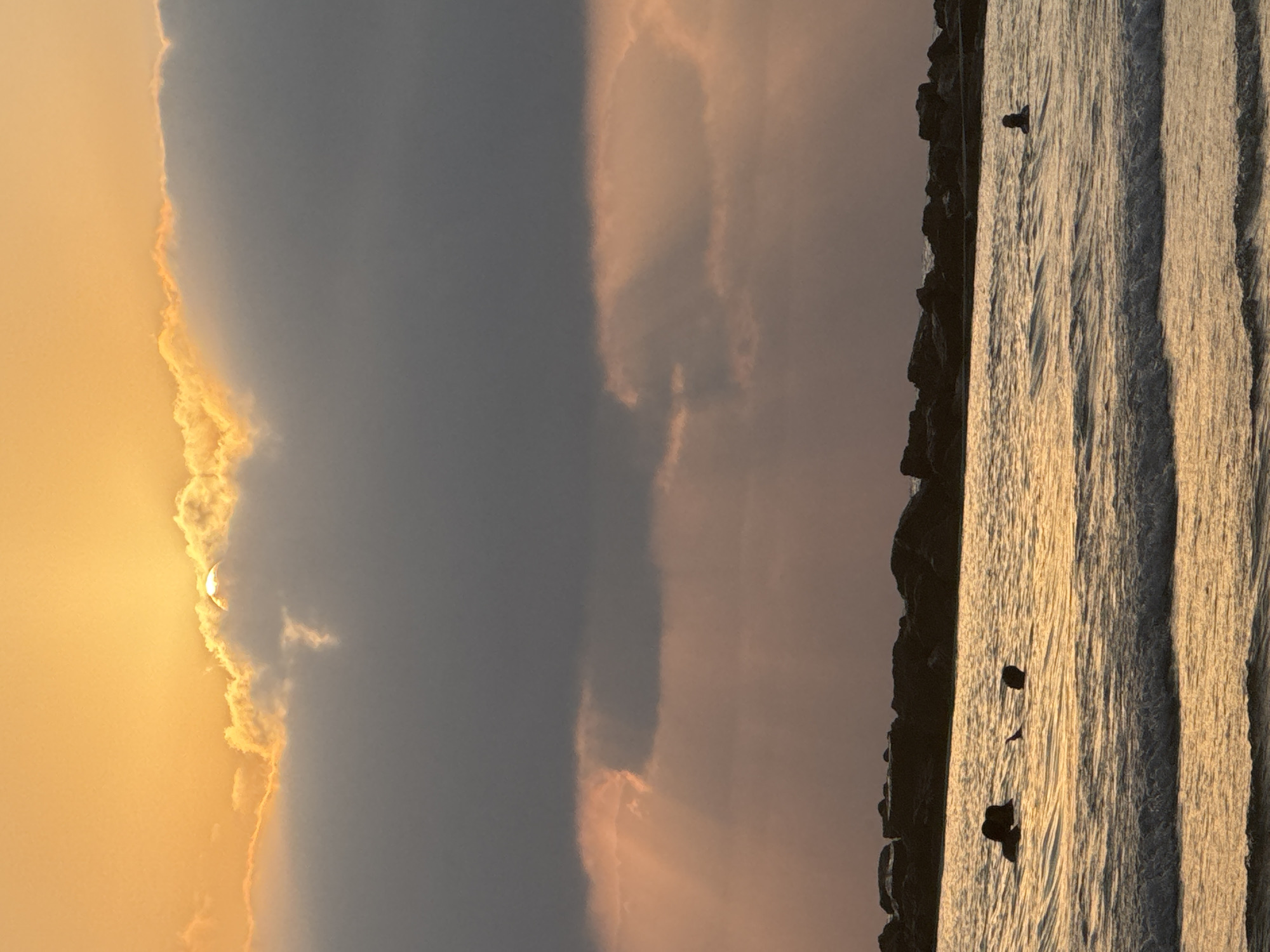 This is a vertical, eye-level, full shot of the ocean at sunset in Netanya, Israel, likely from a beach. The sky is a gradient of orange and yellow at the top, with thick, dark gray clouds obscuring the sun, allowing only a sliver of its light to break through. Rays of sunlight are visible, tinged with pink and orange, radiating downwards through the clouds.
In the middle ground, a line of dark, jagged rocks forms a breakwater or natural barrier along the shoreline. The ocean in the foreground is calm, with gentle waves breaking and creating white foam. The water is reflective, mirroring the colors of the sky.
Several people are visible in the water. To the left, three individuals appear to be swimming or wading. In the center, a person's head and shoulders are visible above the water, with a dark object, possibly a boogie board or a surfboard, nearby. Further to the right, another person's head is visible above the water. The overall scene is serene and captures the beauty of a coastal sunset.