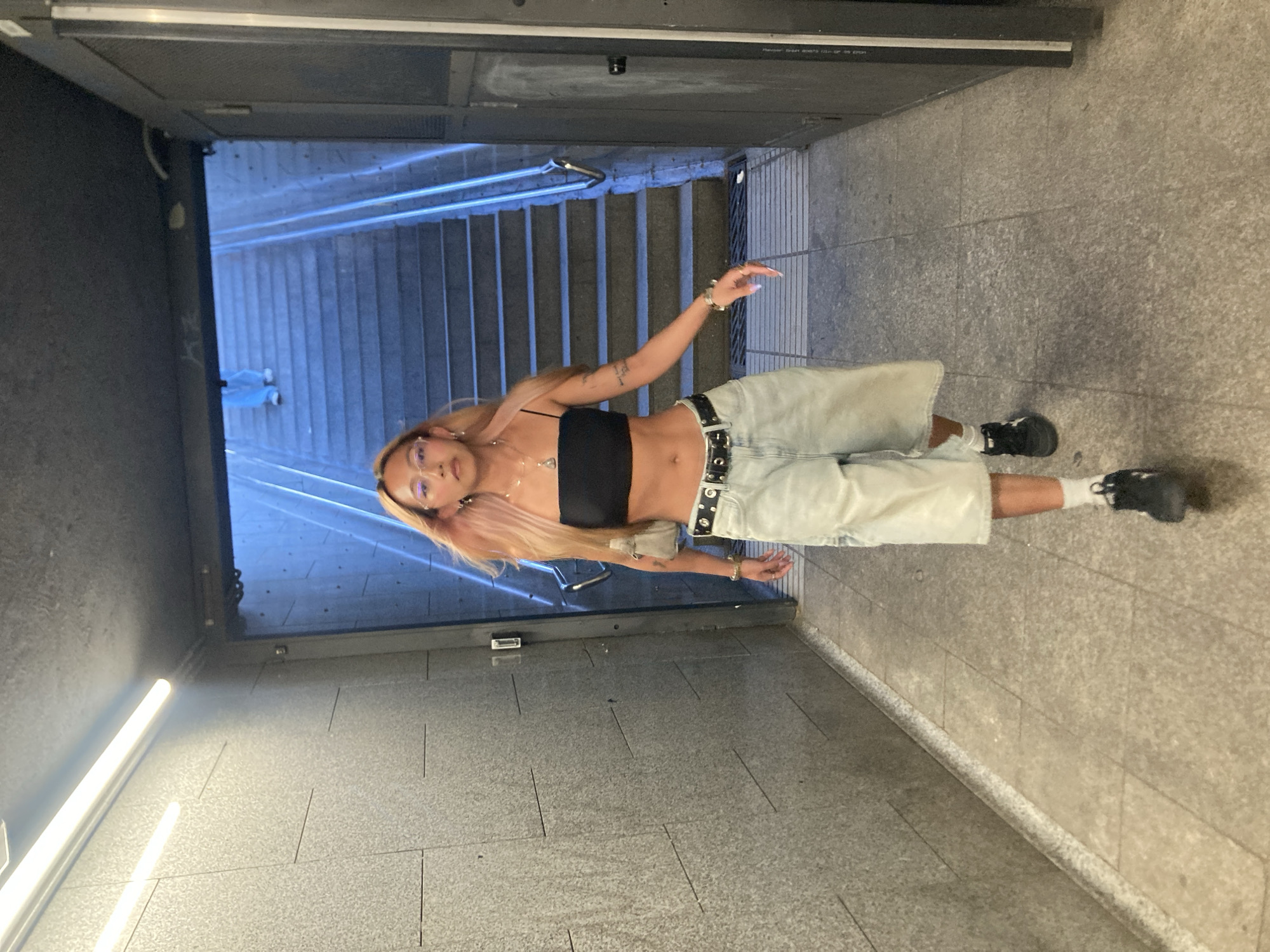 A young woman with blonde hair and glasses walks down a tiled hallway towards a set of stairs. She is wearing a black tube top, light blue baggy shorts, a black belt, white socks, and black sneakers. She has a small purse slung over her shoulder and is wearing jewelry. The hallway has textured grey walls and a tiled floor. The stairs are made of concrete and are illuminated by bright blue lights. A person in blue pants can be seen walking up the stairs in the background. The image appears to be taken in a public underpass or metro station.