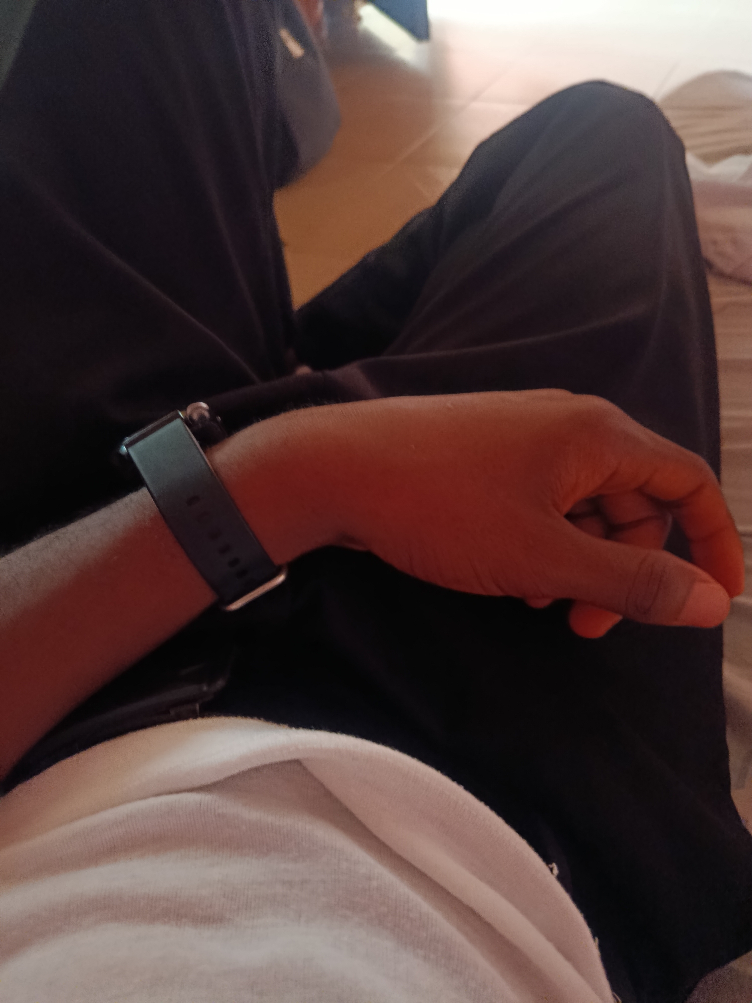 The image shows a close-up of a person's hand and arm wearing a black fitness tracker, resting on dark clothing. The background consists of light-colored fabric and what appears to be wooden flooring. There is no visible indication of the location being in Ukpor, Nigeria. The scene is indoors, with warm lighting. The individual's skin tone suggests they are of African descent. The focus is on the hand, wrist, and the fitness tracker.