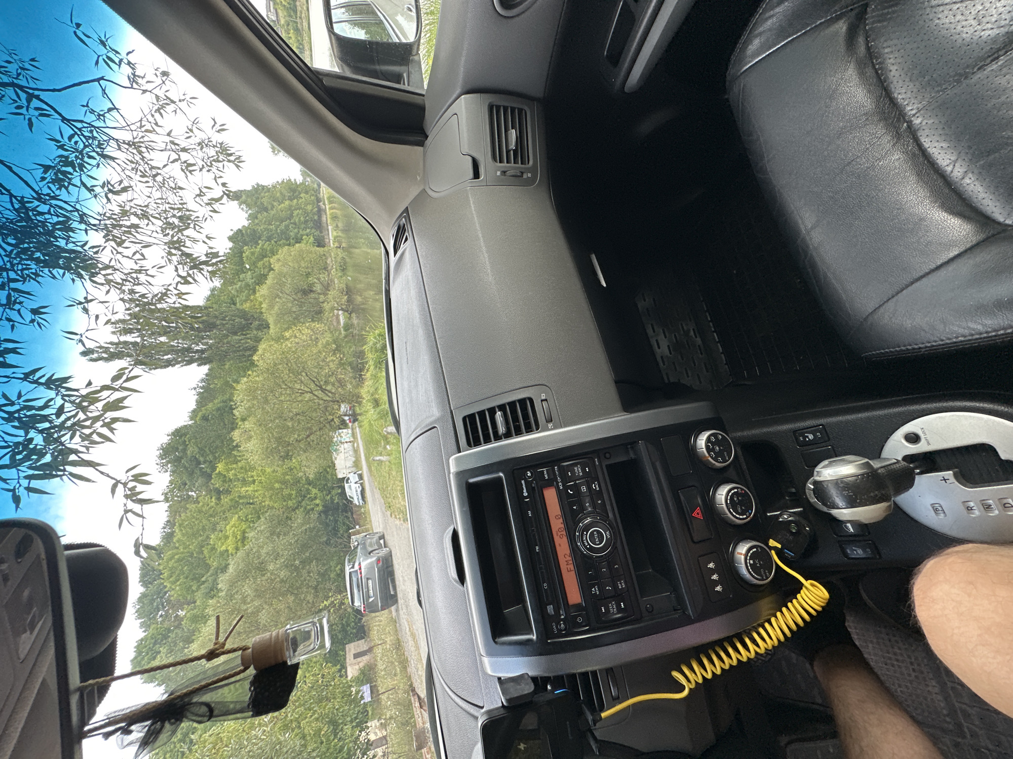 The image shows the interior of a car, likely a Nissan X-Trail, parked on a dirt road in the village of Makiv, Ukraine. Through the windshield, a body of water, likely a river or lake, is visible, bordered by a lush green forest. A grey SUV and a white car are parked further down the road, and several people are walking near the water's edge. The car's dashboard features a multimedia system, climate controls, and a gear shifter. A car air freshener hangs from the rearview mirror, and a coiled yellow cable is plugged into the dashboard. The overall impression is of a peaceful, rural setting on a clear day.