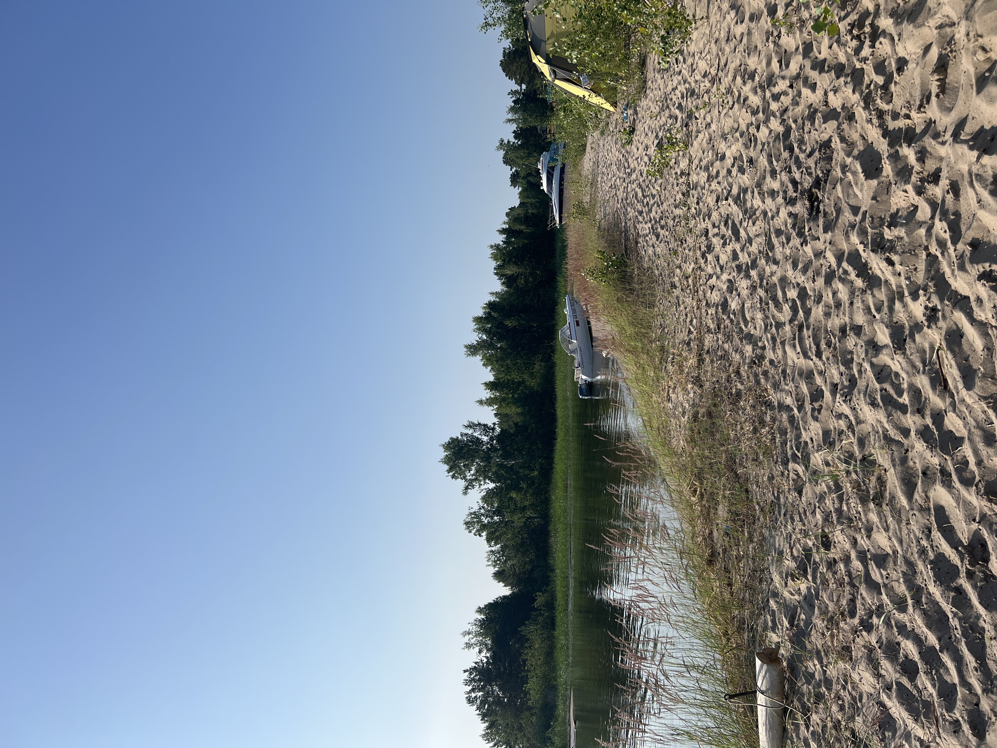 The image depicts a sandy beach area next to a body of water, likely a lake or river, under a clear blue sky. The foreground is dominated by sand with footprints and some sparse vegetation. To the left, tall grasses and reeds grow along the water's edge. In the water, there are two white boats docked near the shore. Further inland, a dense line of green trees forms a backdrop. On the right side of the beach, a yellow and grey tent is set up amidst some bushes. The overall scene suggests a recreational area, possibly a campsite or a spot for boating and relaxation. The location is described as Borovoe, Russia.