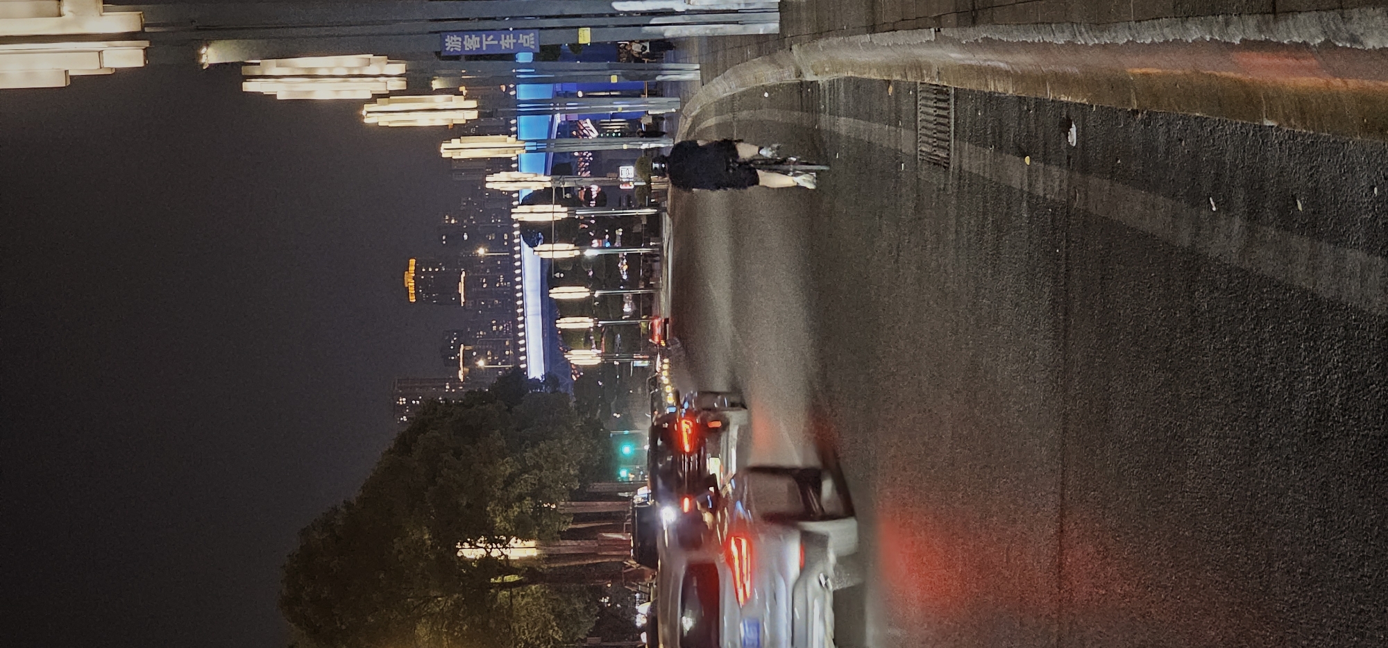 The image depicts a nighttime scene in 江北城街道, China, with a cyclist riding on a city street. Cars with lit headlights and taillights are traveling on the road, indicating traffic. Buildings and illuminated decorative lights are visible in the background, suggesting an urban setting at night.