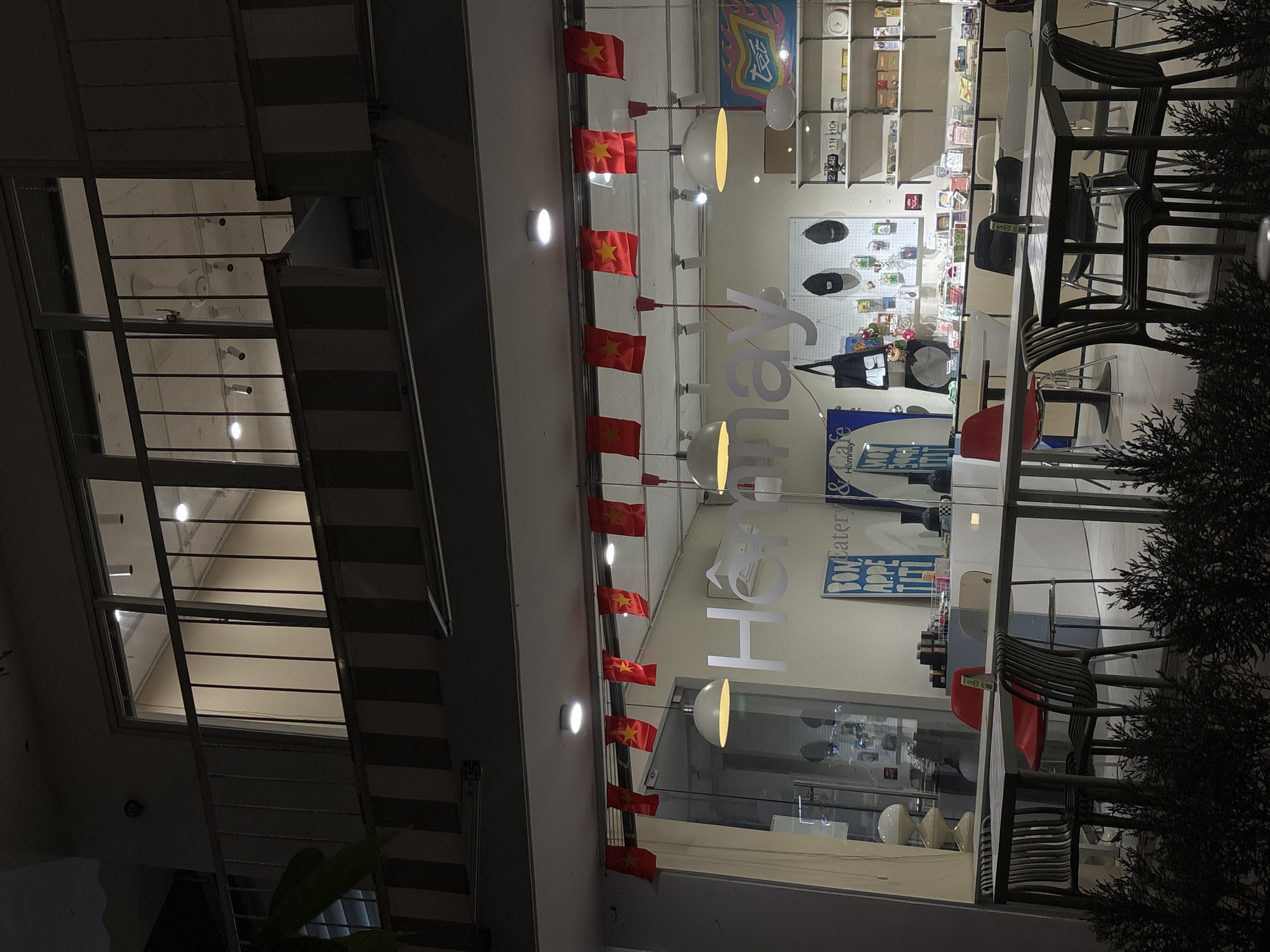 Here is a detailed description of the image:
The image shows an exterior view of a building at night. The facade is decorated with a string of small Vietnamese flags. The building appears to house a shop or cafe called "Honney." The interior of the shop/cafe is visible through the window, displaying shelves of merchandise, and tables with chairs. A plant is partially visible on the right side of the image, and there is a window with blinds to the left of the cafe. The lighting suggests it is evening or night.