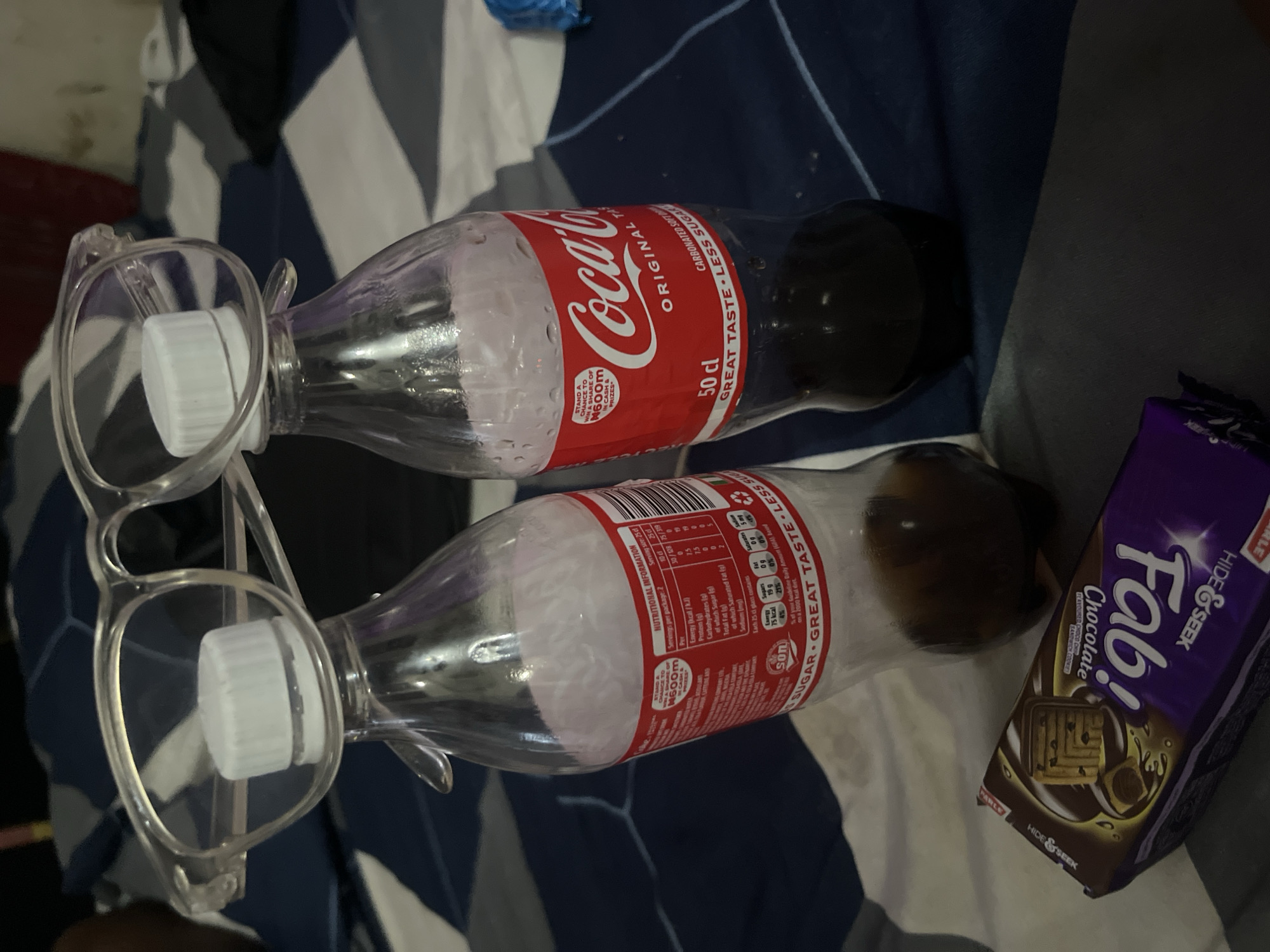 The image shows two partially empty 50 cl Coca-Cola bottles lying on a bed with a white and blue patterned sheet. A pair of transparent-framed glasses rests on the caps of the bottles. Next to one of the bottles is a purple package of "Fab! Chocolate Hide & Seek" cookies. The bottles have red and white Coca-Cola labels with nutritional information. The image provides no information about the location being Lokoja, Nigeria. No people or actions are depicted. The environment appears to be a dimly lit room.