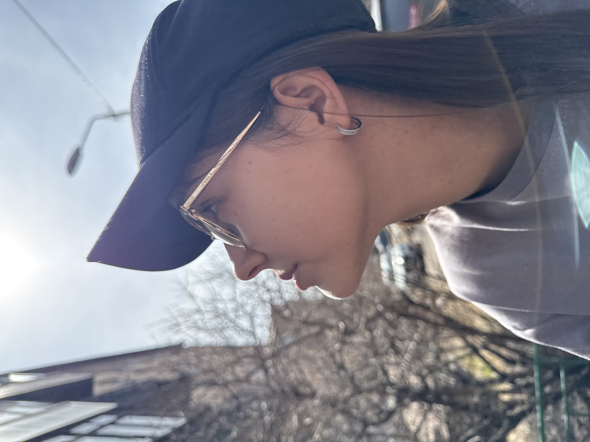A low-angle, medium shot depicts a woman, presumably in Krasnoyarsk, Russia based on image context, facing downwards and to the left. She wears a dark baseball cap, brown hair, and eyeglasses with gold frames; the brand "FURIA" is visible on the temple of the glasses. She also has a small, round earring in her left ear. She is wearing a light grey shirt. Below her, blurred, is foliage and a building. Above her, there is a street lamp and sky. The sun is bright on the left side of the image.