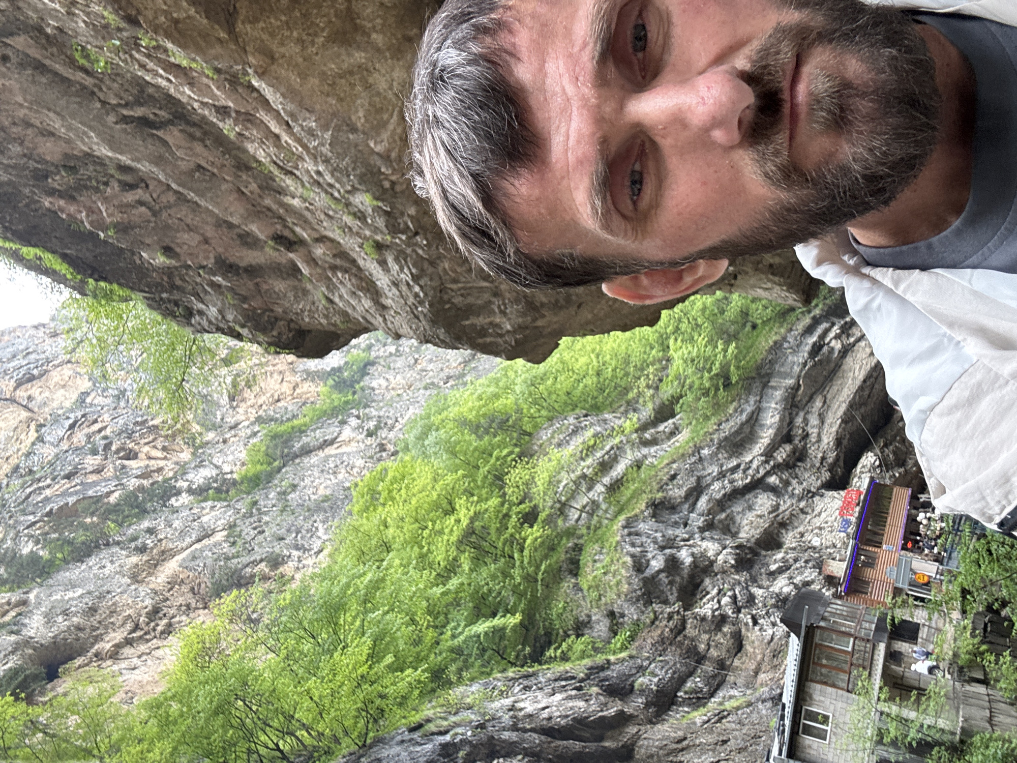 The image shows a man with a beard and graying hair taking a selfie. In the background is a rocky cliff face covered with green trees and vegetation. There is also a building nestled in the cliffside. The specific location in the city of Unknown, Unknown is not identifiable from the image.
