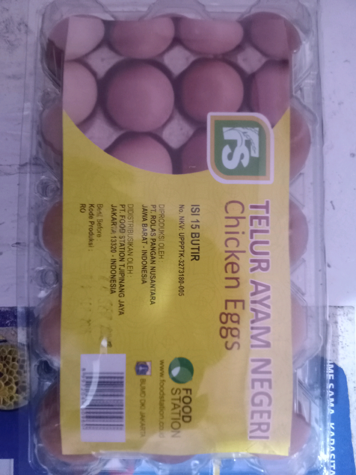 The image shows a package of 15 chicken eggs. The packaging is clear plastic with a yellow label. Text on the label indicates the product is "TELUR AYAM NEGERI" which translates to "DOMESTIC CHICKEN EGGS". The label also includes information about distribution and production. It states "DIDISTRIBUSIKAN OLEH: PT. FOOD STATION TJUIPINANG JAYA JAKARTA 13320 - INDONESIA". This indicates the product is distributed by PT. Food Station Tjuipinang Jaya, located in Jakarta 13320, Indonesia. The text also lists "JAWA BARAT - INDONESIA" under "DIPRODUKSI OLEH: PT. ROLAS PANGAN NUSANTARA". The location of East Jakarta, Indonesia is not explicitly mentioned, but "JAKARTA" is present. The eggs themselves are light brown and are arranged in two rows within the plastic packaging.