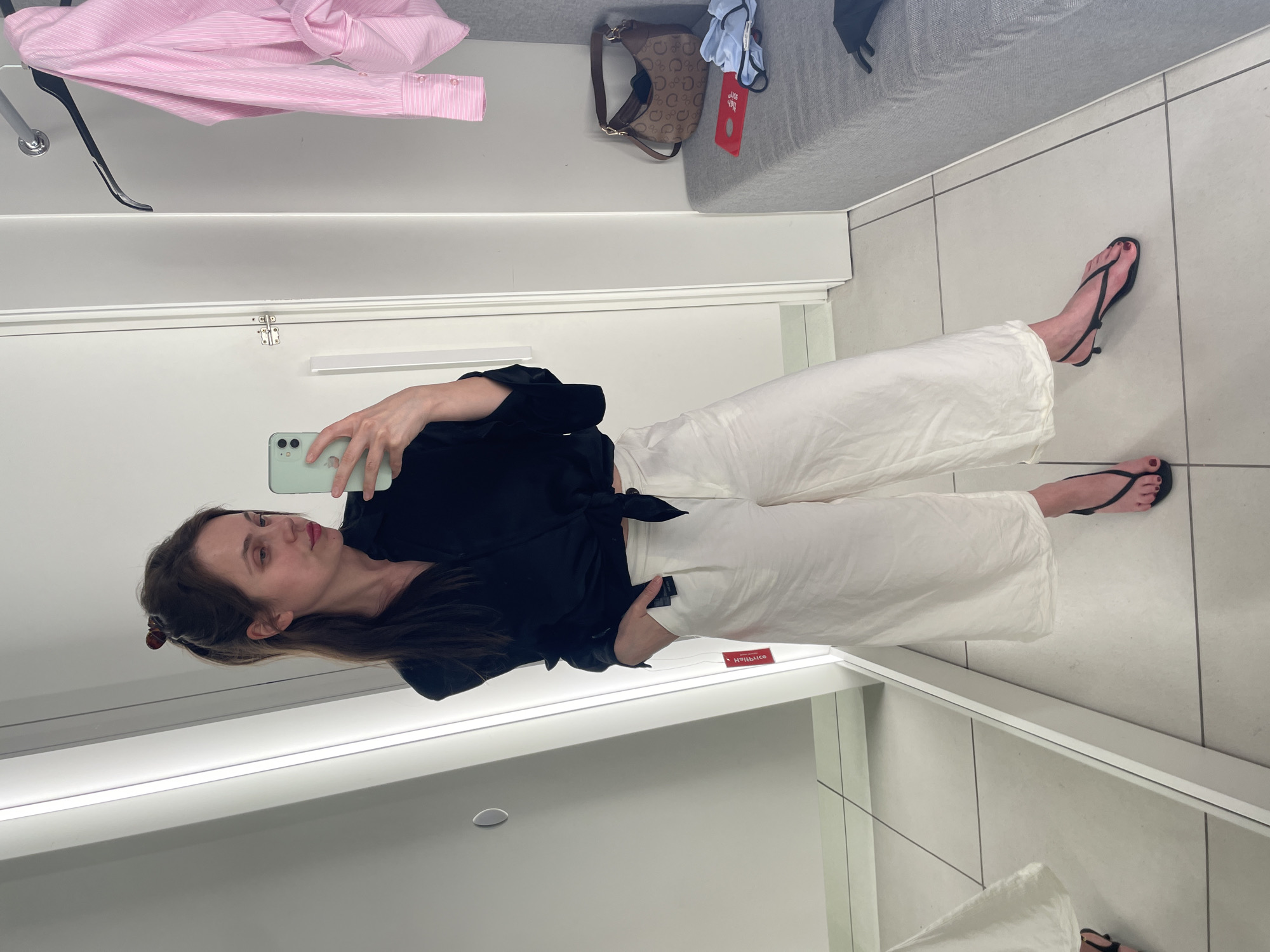 The image shows a woman taking a mirror selfie in a fitting room. She is wearing a black knotted blouse and white culottes. Her hair is pulled back in a ponytail with a clip. She is wearing black thong sandals with a small heel. In the background, there is a pink pinstripe shirt hanging on a rack, a brown patterned handbag on a bench, and a red tag lying on the bench. The fitting room has white walls and a grey bench. The floor is tiled. The location is not specified as Vecsés, Hungary.