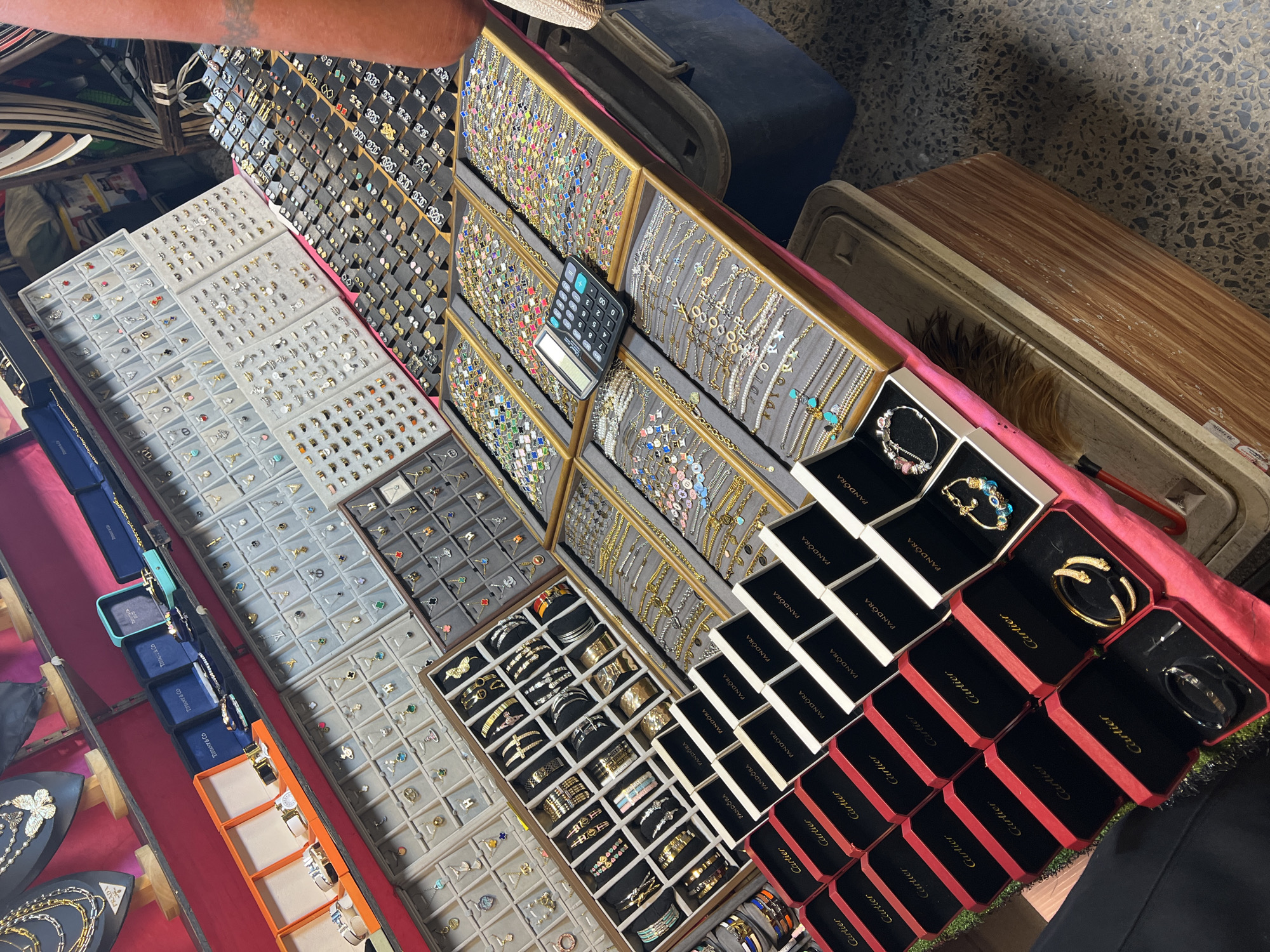 Captured in Baan Bo Phut, Thailand, the image reveals a display of jewelry. The scene is dominated by various trays and boxes containing an assortment of rings, bracelets, necklaces, and earrings. Brands such as Cartier, Pandora, and Tiffany & Co. are clearly visible on the packaging of some items. A calculator rests on one of the jewelry trays. A person's arm is visible in the upper-left corner. The jewelry display is set up on what appears to be a table or counter.