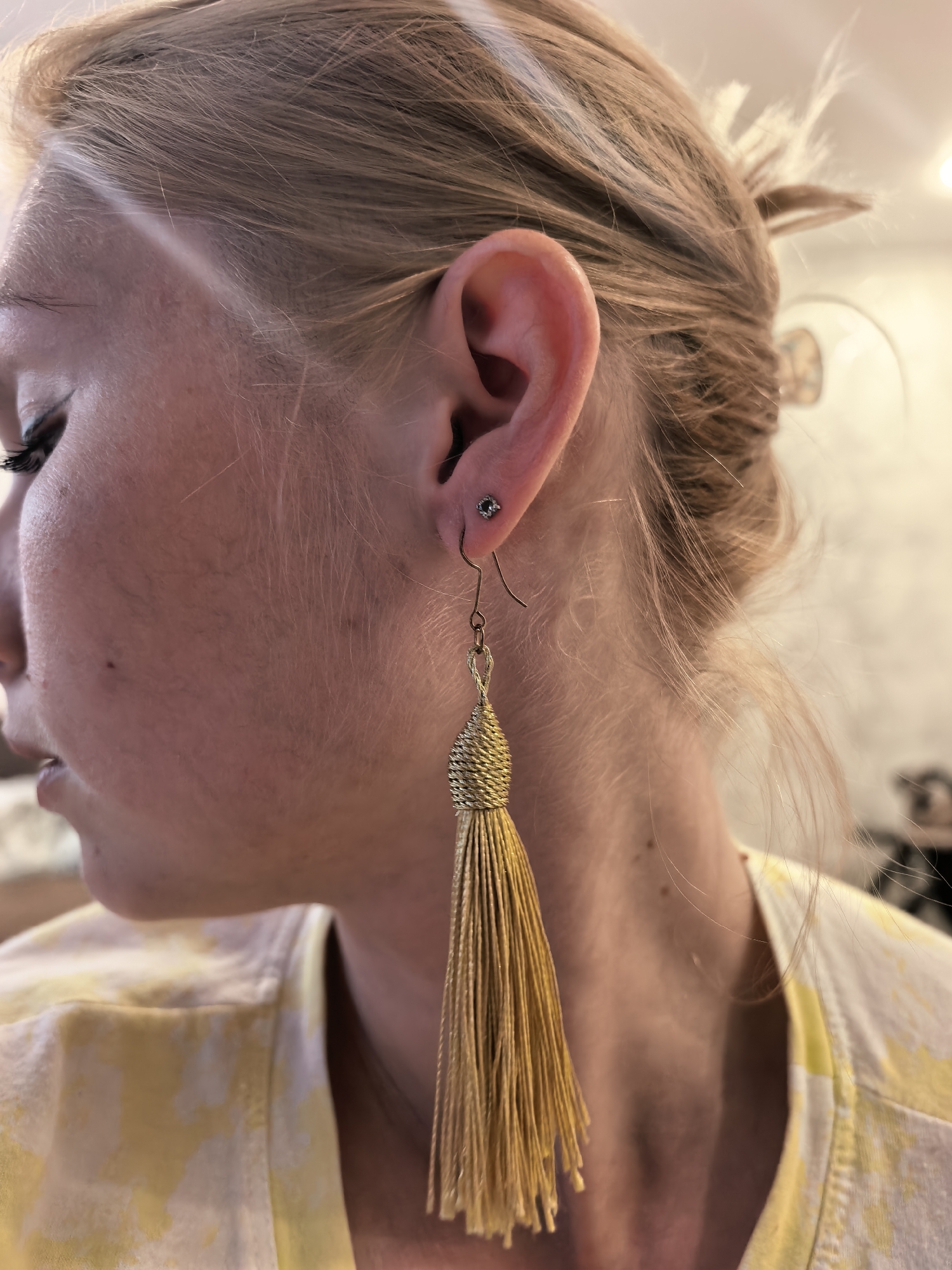 The image is a close-up, side-view portrait of a woman's head and upper body. The woman has fair skin, light blonde hair pulled back, and is wearing a yellow and white tie-dye shirt. She has a small stud earring in her left ear and a long, gold tassel earring hanging from it. The background is blurred, suggesting an indoor setting with soft lighting. The image is taken at close range, focusing on the details of her face, ear, and jewelry. There is no identifiable information in the image that places it in Sterlitamak, Russia.