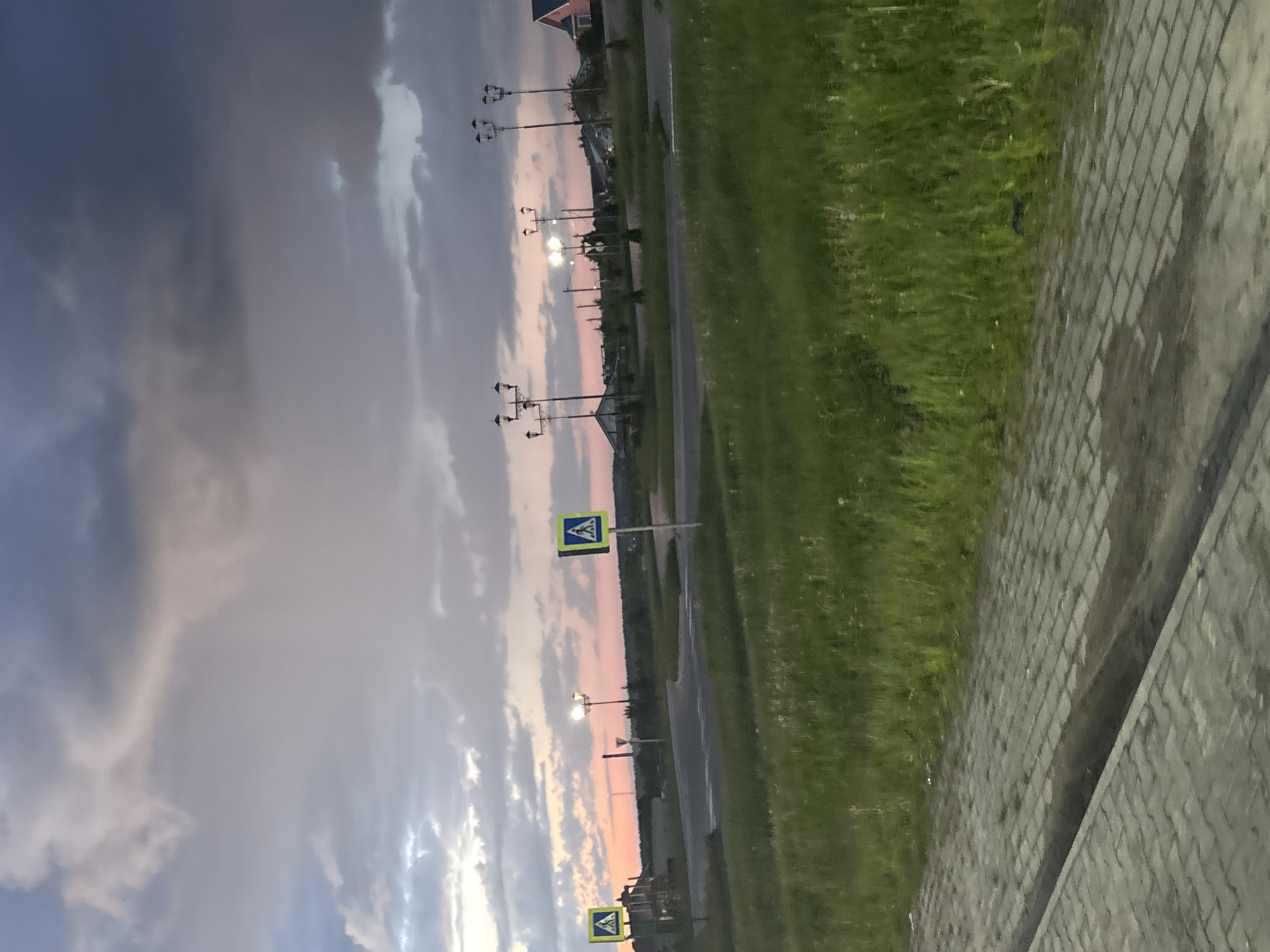 The image captures an outdoor scene at what appears to be either sunrise or sunset, given the soft, warm hues of orange and pink peeking through a predominantly cloudy sky. The photograph is oriented vertically, but depicts a horizontal landscape, suggesting it was taken with the camera held sideways.
The upper portion of the image is dominated by the sky, filled with a mix of dark, heavy clouds and lighter, wispy formations. A gradient of colors, from deep grey-blue on the left to pale pink and orange near the horizon line on the right, signifies the twilight hour. Several streetlights are visible, lining what appears to be a road or paved pathway that stretches into the distance. Some of these lights are illuminated, casting a soft glow.
In the mid-ground, two blue square signs with white triangles and black pedestrian symbols are prominently displayed, indicating pedestrian crossings. These signs are mounted on poles, standing tall beside the road. To the right of the road, a strip of vibrant green grass runs parallel to it, suggesting a verge or roadside lawn.
The foreground on the far right features a paved sidewalk or walkway made of interlocking grey bricks or pavers. This textured surface occupies the lower right quadrant of the image. In the distant background, faint outlines of low-rise buildings or structures can be discerned against the fading light. No people or vehicles are visible, lending a quiet, somewhat serene atmosphere to the scene. There is no discernible text on any objects.