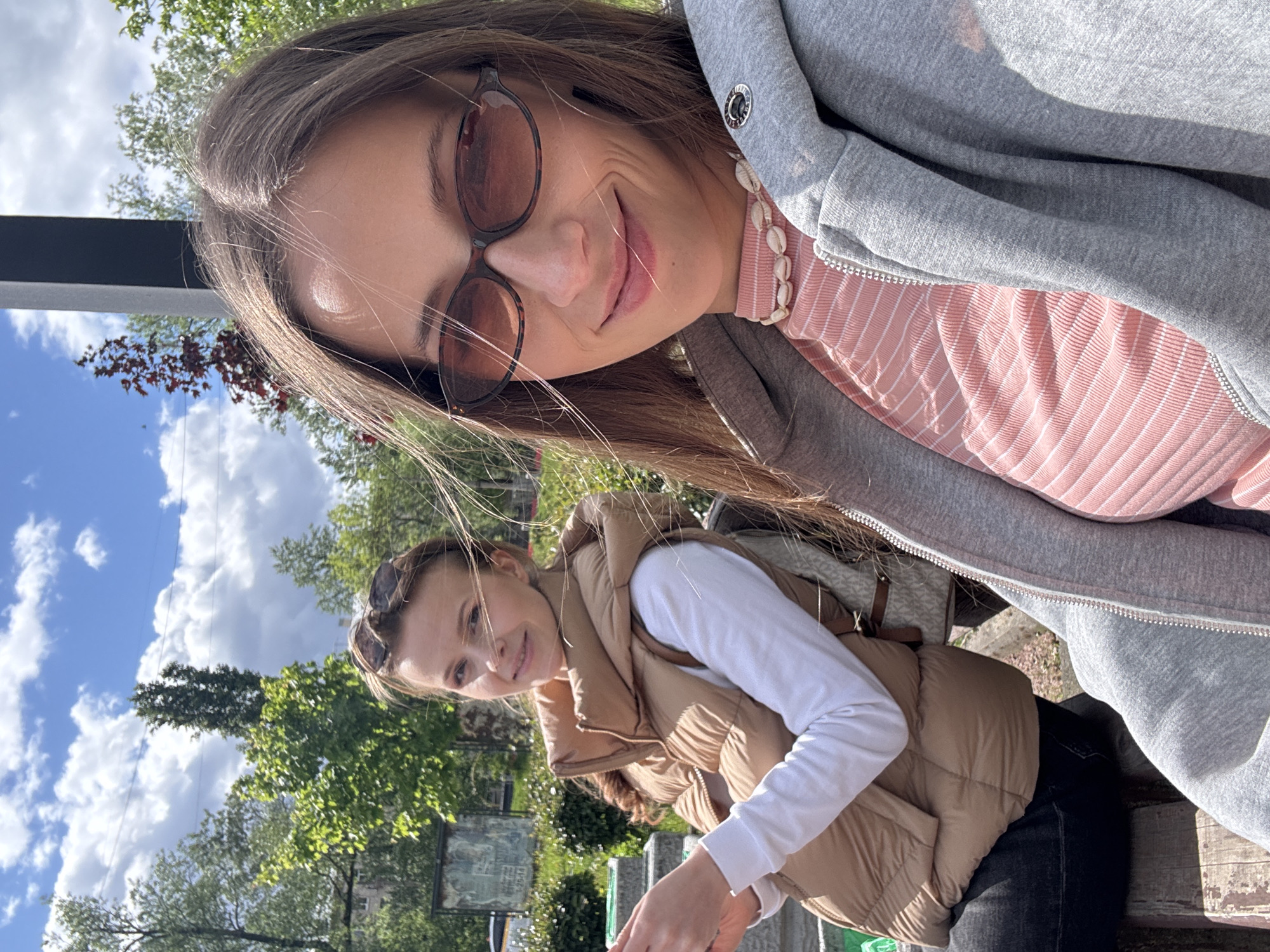 Two women are posing for a selfie outdoors on a sunny day. The woman in the foreground wears brown sunglasses, a gray hooded jacket over a pink and white striped shirt, and a shell necklace. The woman in the background wears a tan puffer vest over a white long-sleeved shirt. They are sitting on a bench. Trees and a cloudy blue sky are visible. Based on visual cues like the stone structures, greenery, and public display signs, the location could possibly be a park or garden within the city of Saint Petersburg, Russia.