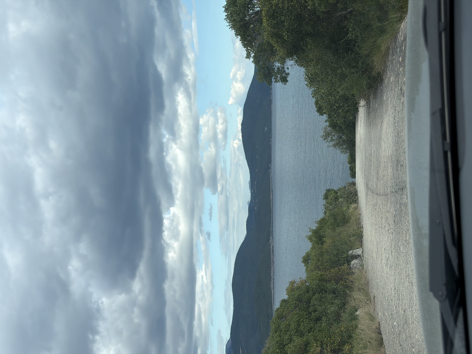 The image depicts a coastal road scene near Radovići, Montenegro. To the right is a gray asphalt road, partly obscured by a gray vehicle component. On the right side of the road are green bushes and trees. Beyond the trees, a gray body of water stretches out, flanked by a dark mountain range in the middle ground. In the sky, a mix of dark gray storm clouds and patches of blue sky with white clouds are present. The viewpoint suggests the photo was taken from inside a vehicle traveling along the road.