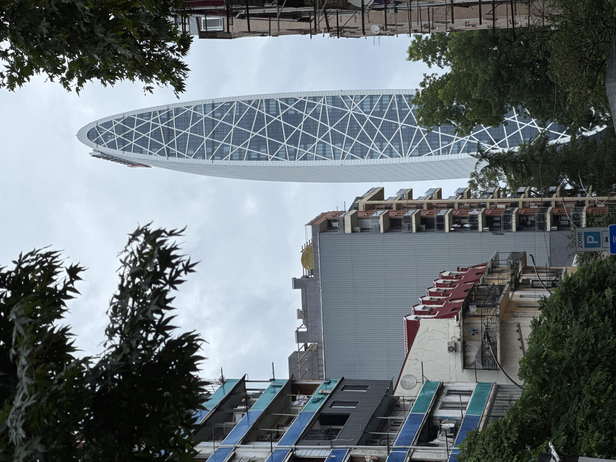 The image depicts a view of Batumi, Georgia, featuring a striking modern skyscraper with a distinctive geometric facade, likely the Hilton Batumi, set against a cloudy sky. In the foreground, several older, multi-story residential buildings are visible, characterized by balconies, exposed utilities, and some visible laundry hanging out. Lush green trees partially obscure the view of the buildings and the skyscraper, suggesting an urban environment with pockets of nature. A street sign indicating a parking zone is visible in the lower right corner. The overall impression is a juxtaposition of old and new architecture within a city setting.