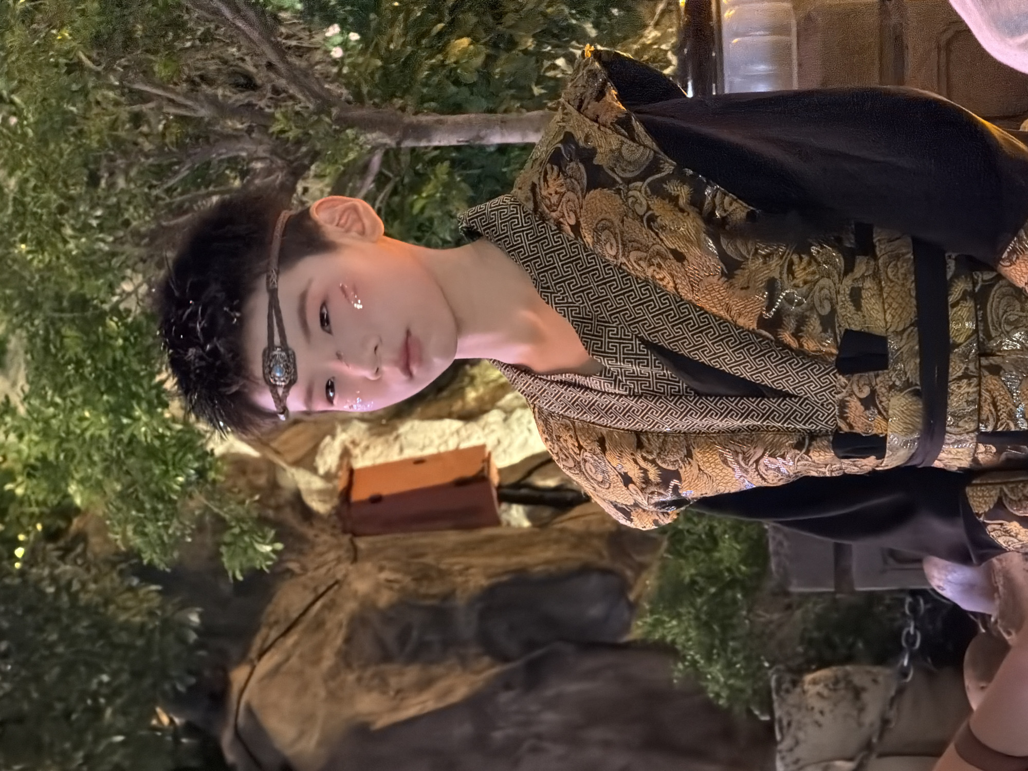 A young boy with dark hair and glitter makeup on his face stands in front of a rocky, shrub-filled backdrop. He is wearing a traditional black garment with a gold, dragon-patterned vest and a woven headband. The setting appears to be an outdoor evening event with decorative lighting visible in the background. The location is identified as Yuzhong District, China.