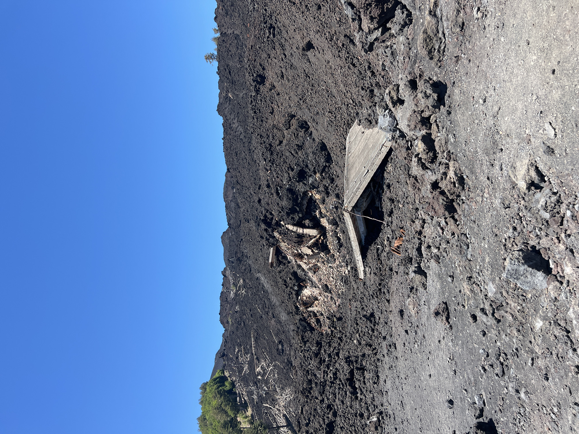 The image depicts a desolate volcanic landscape, likely on Mount Etna near Linguaglossa, Italy. The foreground and midground are dominated by jagged, dark volcanic rock and ash, characteristic of a recent eruption or lava flow. Scattered within this rugged terrain are the remnants of a destroyed structure. A partially collapsed wooden roof lies askew, resting on the volcanic debris. Further back, more substantial structural elements, including concrete and rebar, are visible, indicating a building that has been severely damaged or engulfed. The background shows a sloped hill covered in the same dark volcanic material, with some sparse green vegetation and trees on the upper left side, suggesting the edge of a forest that has been impacted by the volcanic activity. The sky above is a clear, vibrant blue, indicating fair weather. There are no people or animals visible in the scene.