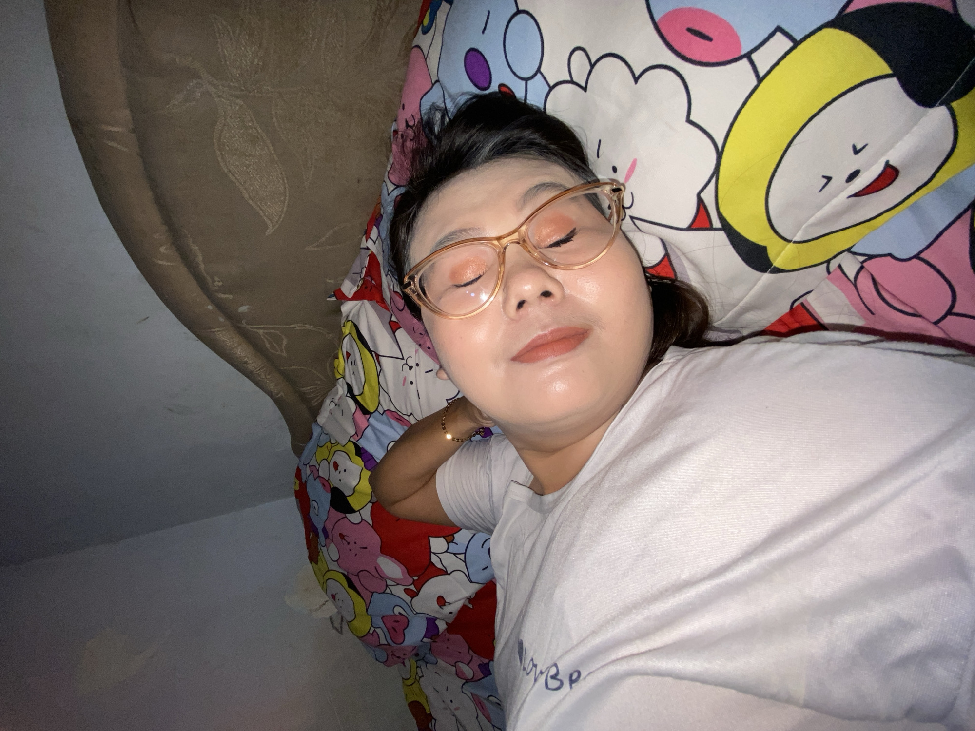A young woman with closed eyes is lying on her back in bed, wearing glasses and a white t-shirt. She is wearing makeup, including eyeshadow and lipstick. The bed is covered with a colorful, cartoon-patterned comforter and pillow. The room appears to be dimly lit, with the bed positioned against a wall. The scene is consistent with a bedroom in a residential setting in Gelam, Indonesia.