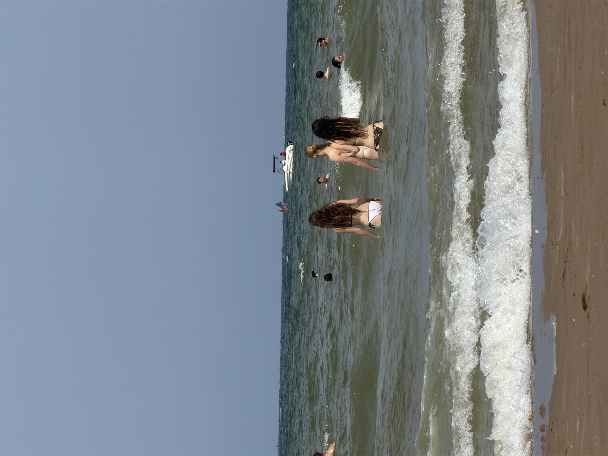 The image shows a sunny day at a beach. Several people are in the water, some swimming and others wading. In the foreground, there are two women in bikinis, with their backs to the camera. One woman has long, dark hair, and the other has blonde hair. In the background, there is a white boat with a few people on it, and a flag flying from a pole. The sky is clear and blue, and the water is a greenish-brown color with moderate waves. The sand in the foreground is damp, indicating that it is close to the water. This scene is consistent with a beach on the Mediterranean coast, potentially in Valencia, Spain, known for its numerous beaches and popular watersports.