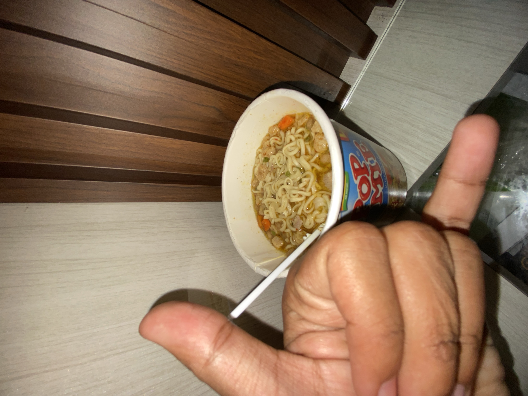 The image shows a cup of instant noodles with a white plastic fork inside, partially obscured by a human hand making a gesture. The cup appears to be on a surface, possibly a table or counter, with a light-colored, wood-grain pattern. A darker, horizontally-paneled material is visible in the background. Based solely on the provided image, no information about the location in Lhokseumawe, Indonesia can be determined.