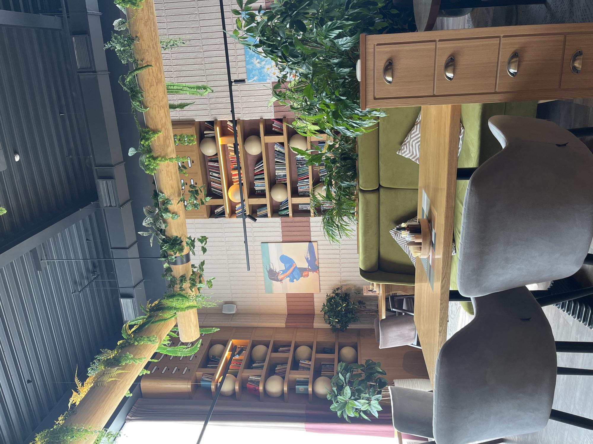 The image depicts the interior of a restaurant or cafe located in Verkhnyaya Pyshma, Russia. The interior design is characterized by a blend of modern and natural elements. A wooden beam adorned with greenery runs across the ceiling, adding a touch of nature. Bookshelves are built into the walls, with decorative spherical objects interspersed among the books. Paintings are mounted on the walls. A green velvet sofa is positioned near a wooden table, and chairs with gray fabric upholstery are placed around the table. A wooden cabinet with drawers stands adjacent to the sofa. The overall environment suggests a relaxed and inviting atmosphere.
