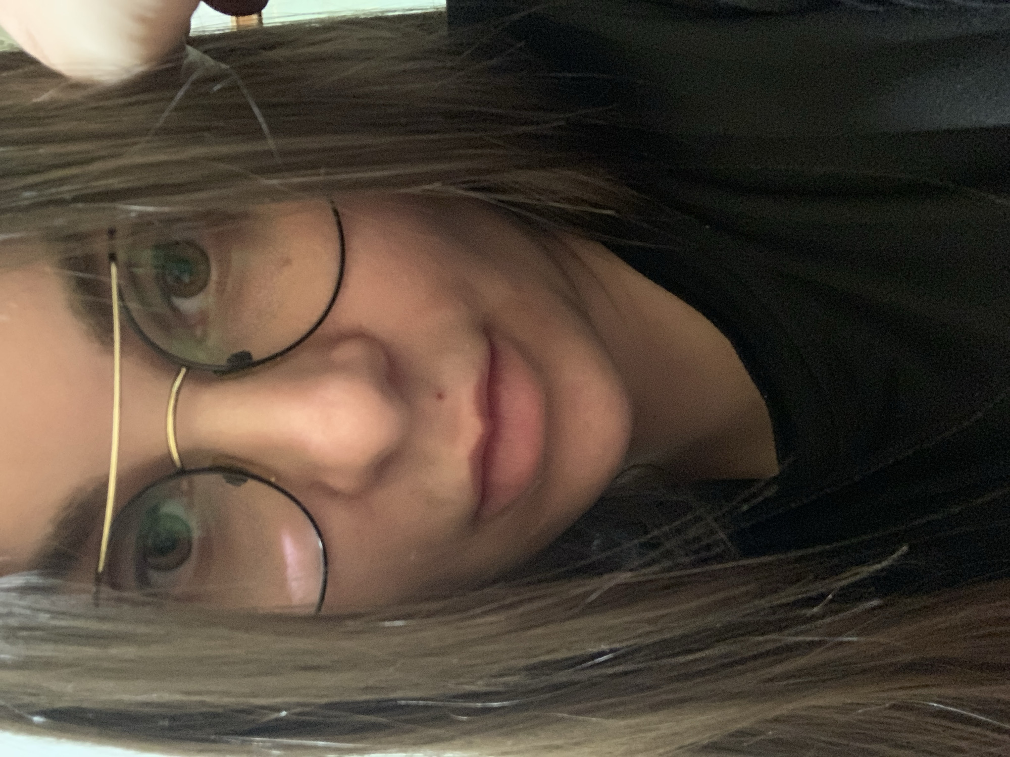A close-up, frontal, waist-up, indoor photograph shows a young woman with light brown hair and light skin. She is wearing large, round, wire-rimmed glasses with gold frames. Her eyes are light brown, and she is looking directly at the camera. She is wearing a black t-shirt or blouse. Her skin is smooth and clear, with a small mole on her cheek just below her left eye and another on her upper lip. Her lips are full and pink. Her hair is long and falls around her face and shoulders. Her right hand is visible in the upper right corner of the image, touching her hair. The background is out of focus and appears to be a plain, light-colored wall.