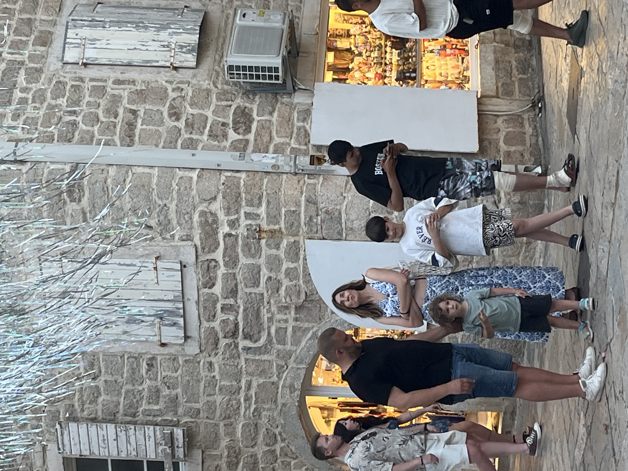 A group of people are walking on a stone-paved street in front of a building with stone walls and wooden shutters. The building has a shop with brightly lit merchandise visible through its window. There are decorative silver streamers hanging from above, possibly for an event or decoration. The people include a man and a woman with two children, as well as other individuals walking past. The scene appears to be in an urban or town setting, likely a pedestrian area. The overall impression is of a casual stroll on a street.