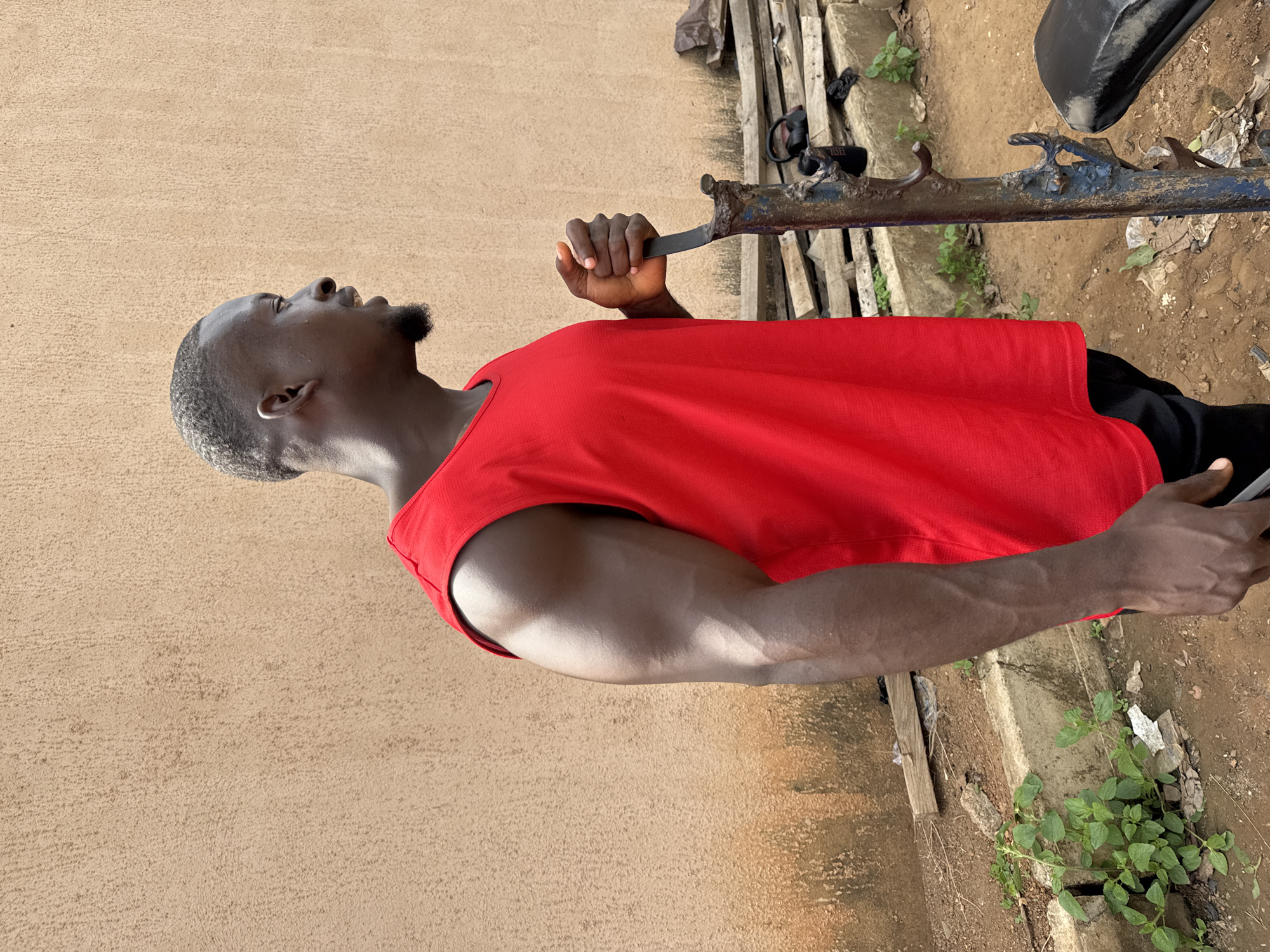 A profile view of a man with dark skin and short black hair stands outdoors, facing right, against a textured tan wall. He is wearing a red sleeveless shirt and black shorts. His right arm is flexed, showcasing developed musculature. He holds a weathered, rusty metal bar vertically with his right hand, his grip tight. In the background, wooden planks are stacked, and green plants grow near a concrete border. The ground is rough dirt.