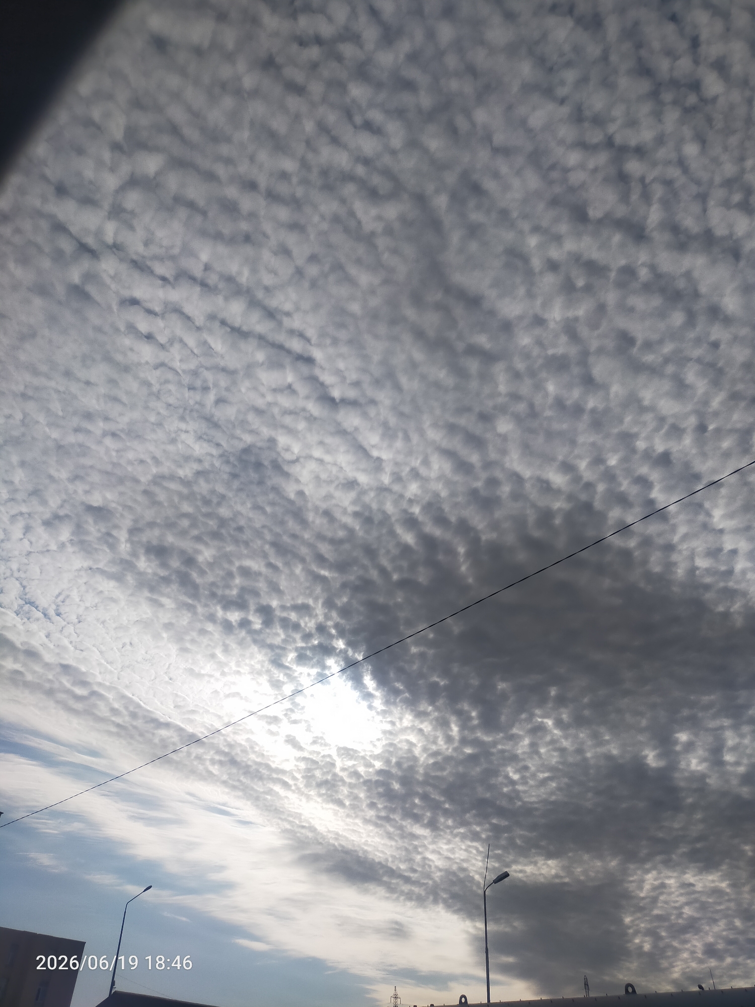 The media file is an outdoor photograph taken from a low angle, looking up at a dramatic sky filled with altocumulus clouds. These clouds are arranged in a textured pattern of small, rounded masses, creating a rippled or mackerel-like effect. The sun is partially obscured by the clouds, casting a bright, diffused light through the gaps, particularly in the upper left quadrant of the image.
The lower portion of the image shows hints of a man-made environment. A dark building or structure is visible on the far left, and a street lamp with a bent pole stands out against the sky. A thin, dark wire stretches diagonally across the sky. The sky transitions from a bright, sunlit area to darker, more ominous clouds towards the right.
The time stamp "2026/06/19 18:46" is visible in the lower left corner, indicating the date and time the photograph was taken. This suggests it is late afternoon or early evening. The weather appears overcast with some bright patches, creating a dynamic and somewhat dramatic atmosphere. The scene is devoid of people. The overall impression is one of natural beauty and the vastness of the sky.