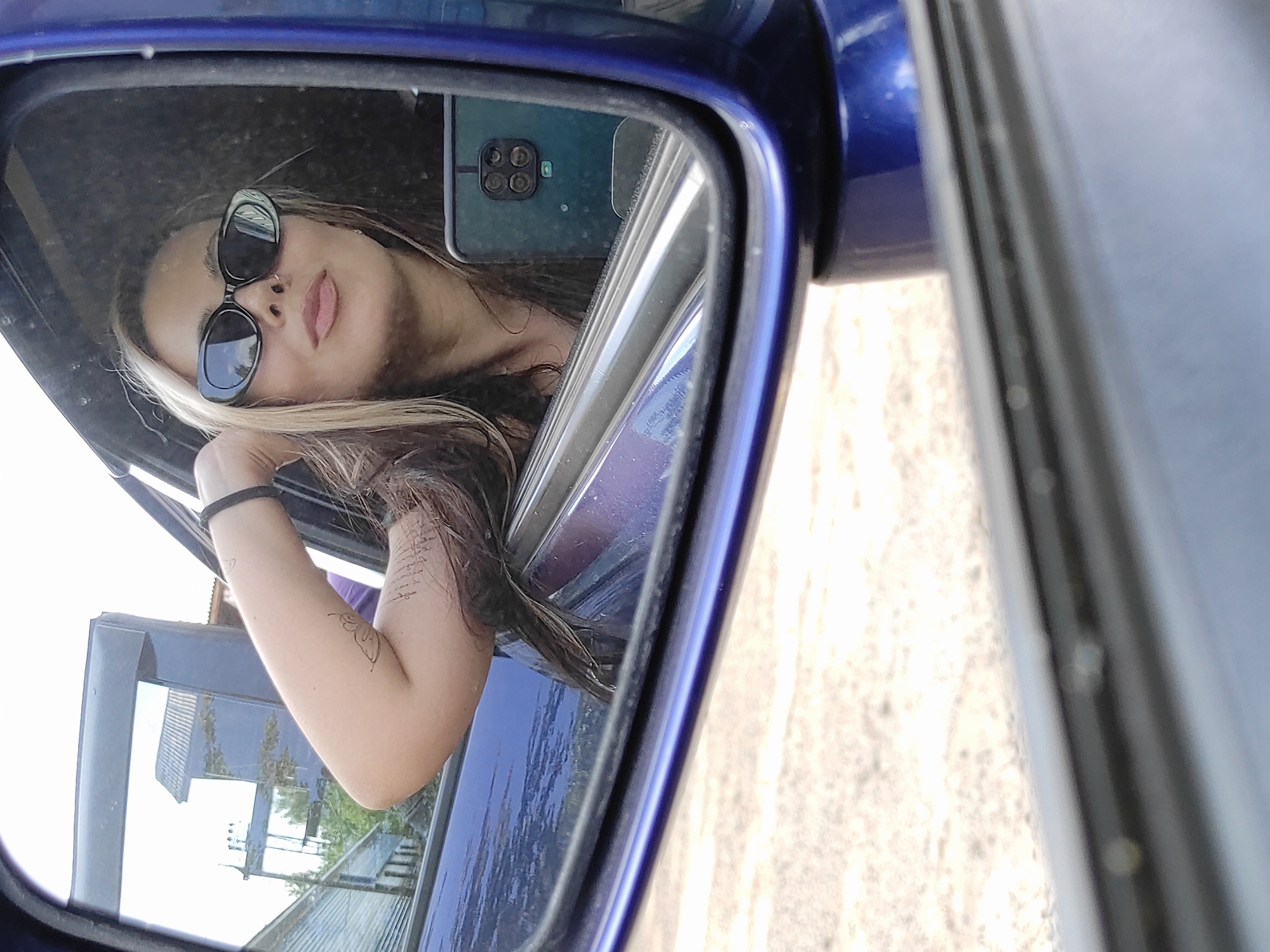 A woman in sunglasses is reflected in a blue car's side mirror. The woman is holding a cell phone with four visible camera lenses. The reflection in the mirror shows her arm and part of the open car door. Outside the car, there's a view of a building and some vegetation. The location is Chaikovsky, Russia.