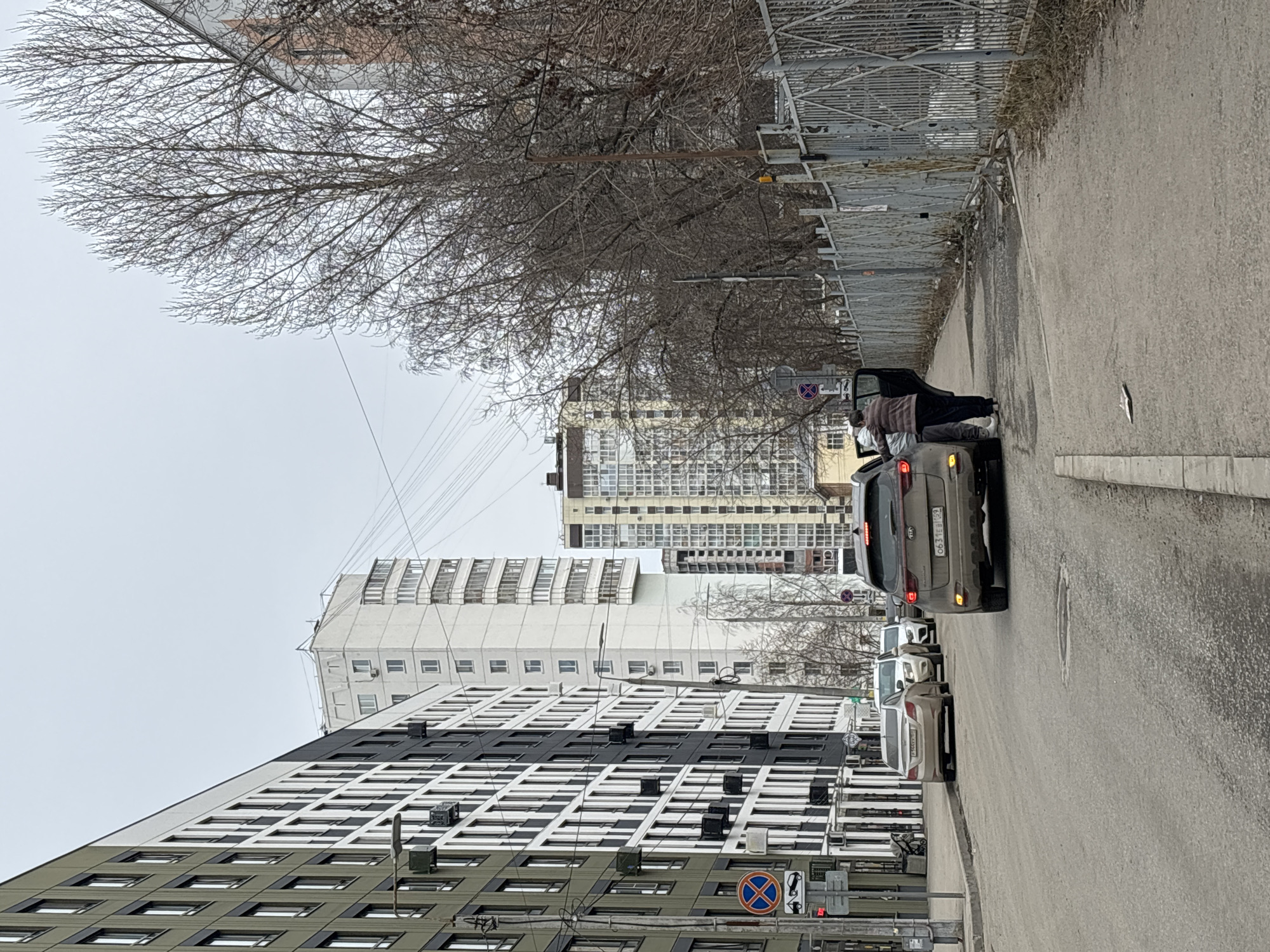 The image depicts a street scene in Perm, Russia. A multi-story building with a modern, grid-like facade of alternating dark and light panels dominates the left side of the frame. In the background, another taller building is visible. An overcast sky casts a muted light over the scene.
On the right side of the frame, a row of parked cars lines the street. A gray Kia SUV is parked with its driver-side door open. Two individuals are standing next to the car, appearing to interact. One person is wearing a striped shirt, dark pants, and white shoes, while the other's attire is partially obscured. The asphalt road is cracked and marked with patches and uneven surfaces.
A metal fence runs along the edge of the sidewalk, with bare trees visible behind it. The branches of the trees are stark against the gray sky. Overhead power lines crisscross above the buildings. Traffic signs, including one with a red circle and a blue cross indicating "No Parking", are present near the buildings.