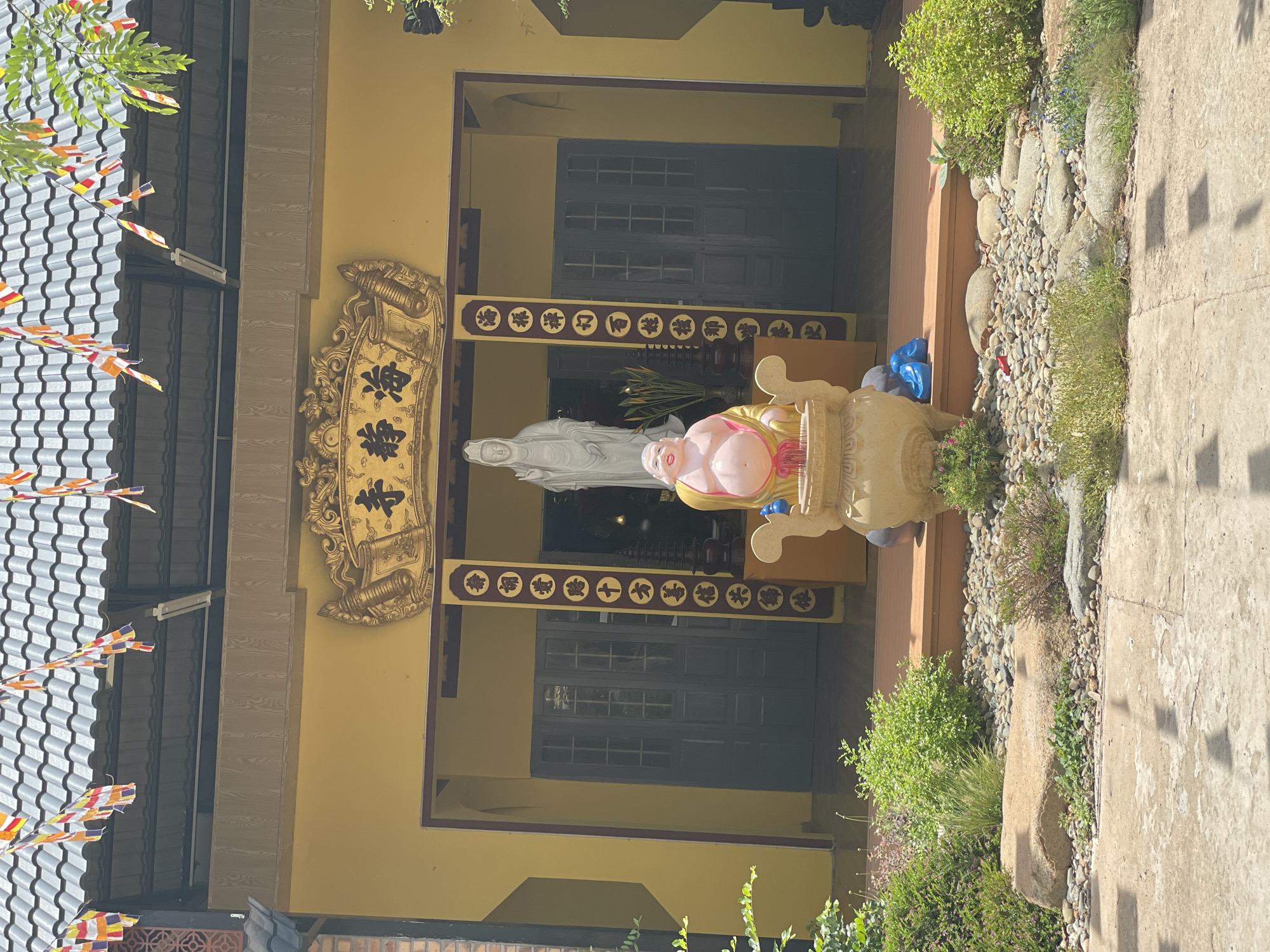 The image shows the exterior of a yellow building, likely a Buddhist temple, in Mũi Né, Vietnam. In front of the building are two statues: a large, white statue of a female figure standing on the right, and a smaller, smiling, pot-bellied statue of a man sitting on the left, in front of the white statue. The pot-bellied statue is placed in front of a large, decorative urn. The building has a tiled roof adorned with prayer flags. Above the entrance, a golden plaque with intricate carvings and Chinese characters is visible. The entrance itself features a pair of dark doors and is flanked by vertical banners with more Chinese characters. The foreground of the image consists of a paved area, with a rock garden featuring various sized stones and small green plants. The overall scene is brightly lit by sunlight, casting shadows on the pavement.