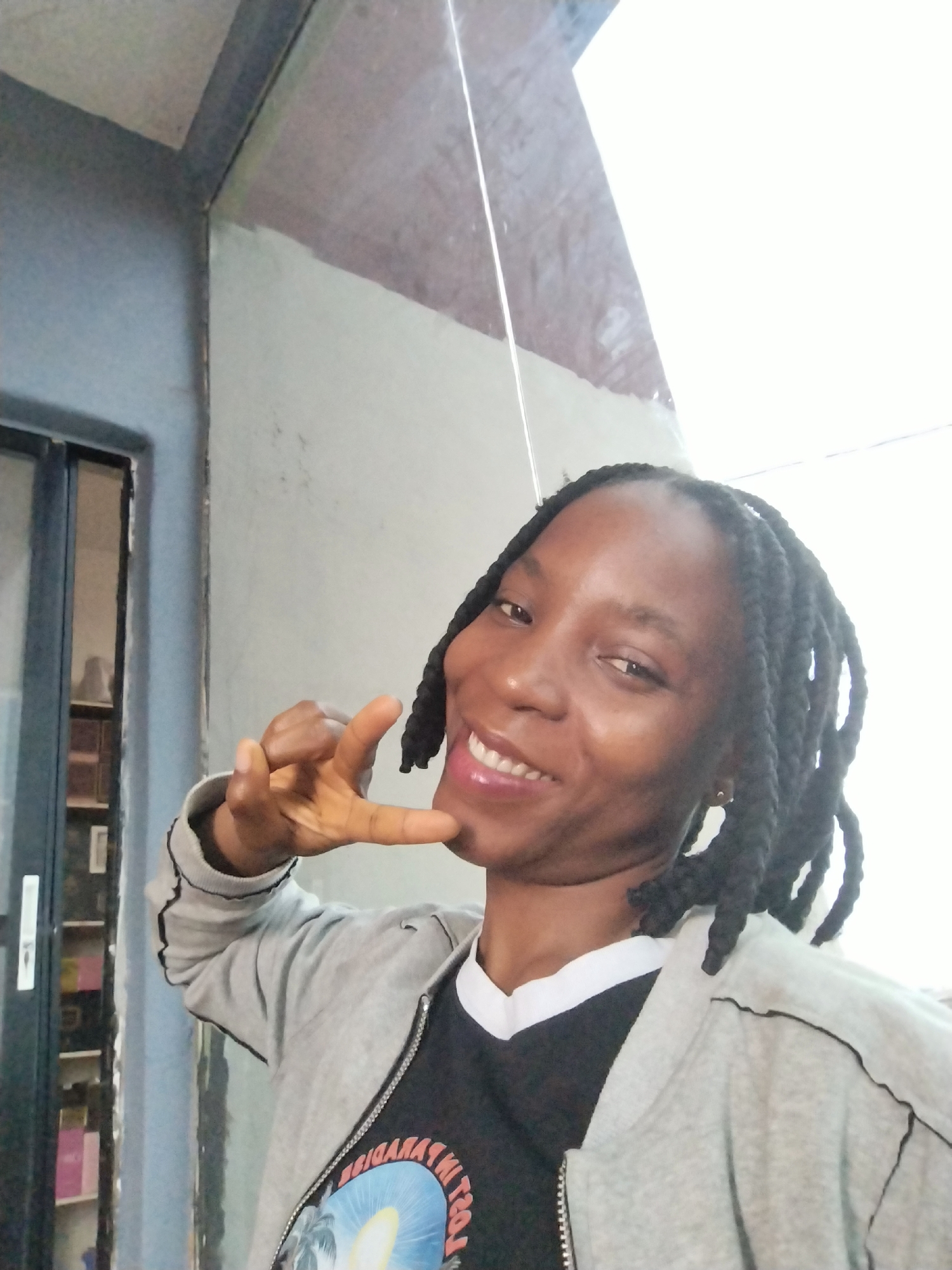 A Black woman is pictured from the chest up, smiling at the camera in an outdoor setting. She has dark braided hair, light brown skin, and is wearing a grey zip-up hoodie over a black t-shirt with a graphic print. Her right hand is raised to her face, with her thumb and index finger touching her chin, and her other fingers extended. The background consists of textured walls, a window or door on the left, and what appears to be a bright, overcast sky. The location is specified as Uselu, Nigeria.