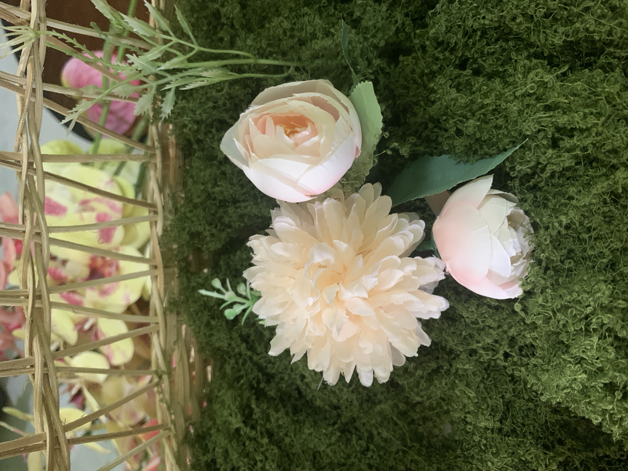 The image depicts a close-up view of artificial flowers and greenery, arranged in a way that suggests a decorative display. In the foreground, there is a bed of textured, moss-like green material, which serves as the base for the arrangement. Three artificial flowers are prominently featured: a large, fluffy, pale peach-colored flower on the left, and two smaller, bud-like pink and white flowers on the right and bottom center. Green artificial leaves are also visible, interspersed with the flowers. In the background, a portion of a woven wicker basket is present, revealing a blurred backdrop of what appears to be more artificial flowers, with colors ranging from yellow to pink. The overall scene evokes a sense of artificial floral decoration. There is no visible indication of its location in Kuala Pembuang, Indonesia.